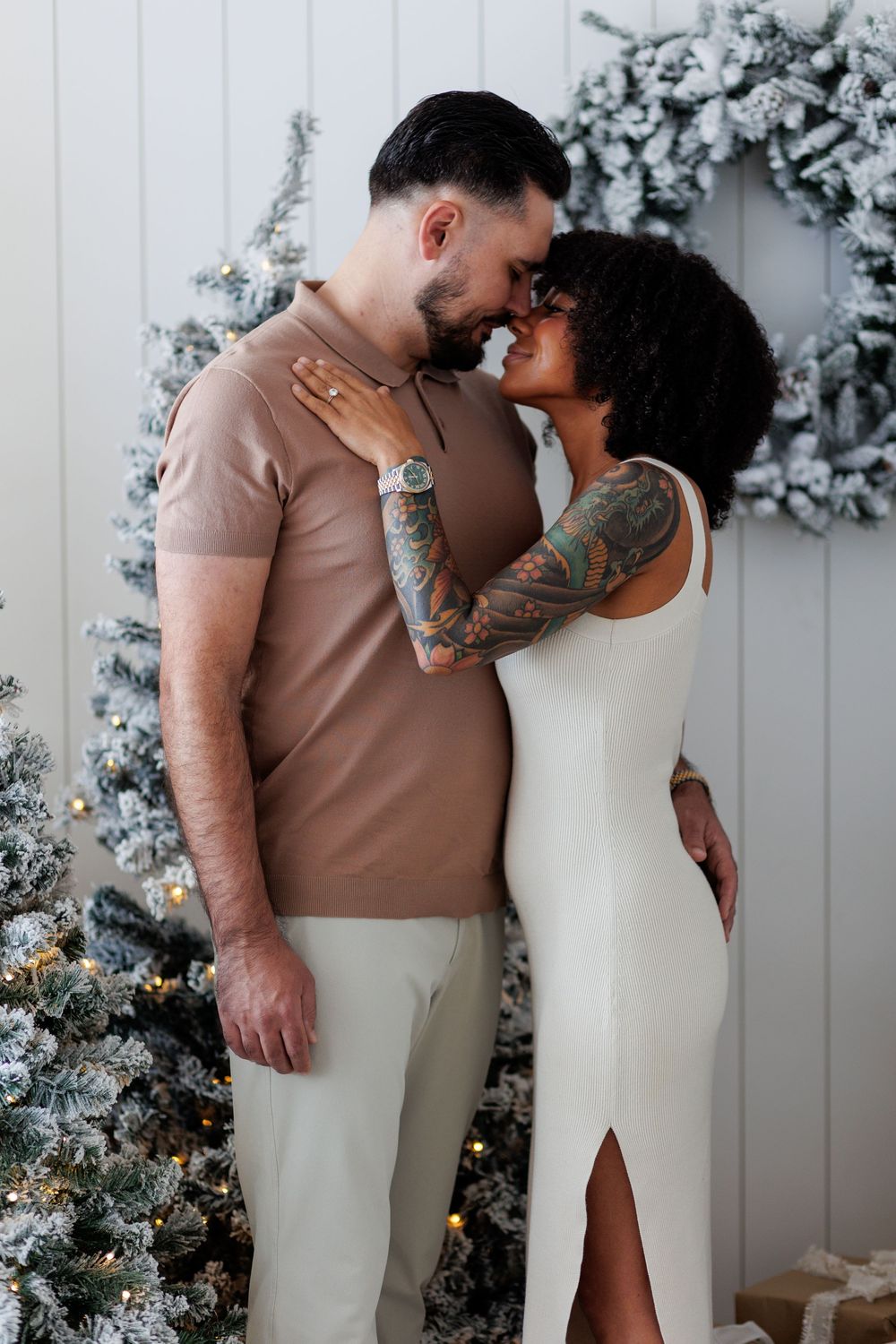 Tattooed interracial couple in neutral outfits sharing a tender moment near frosted Christmas trees.