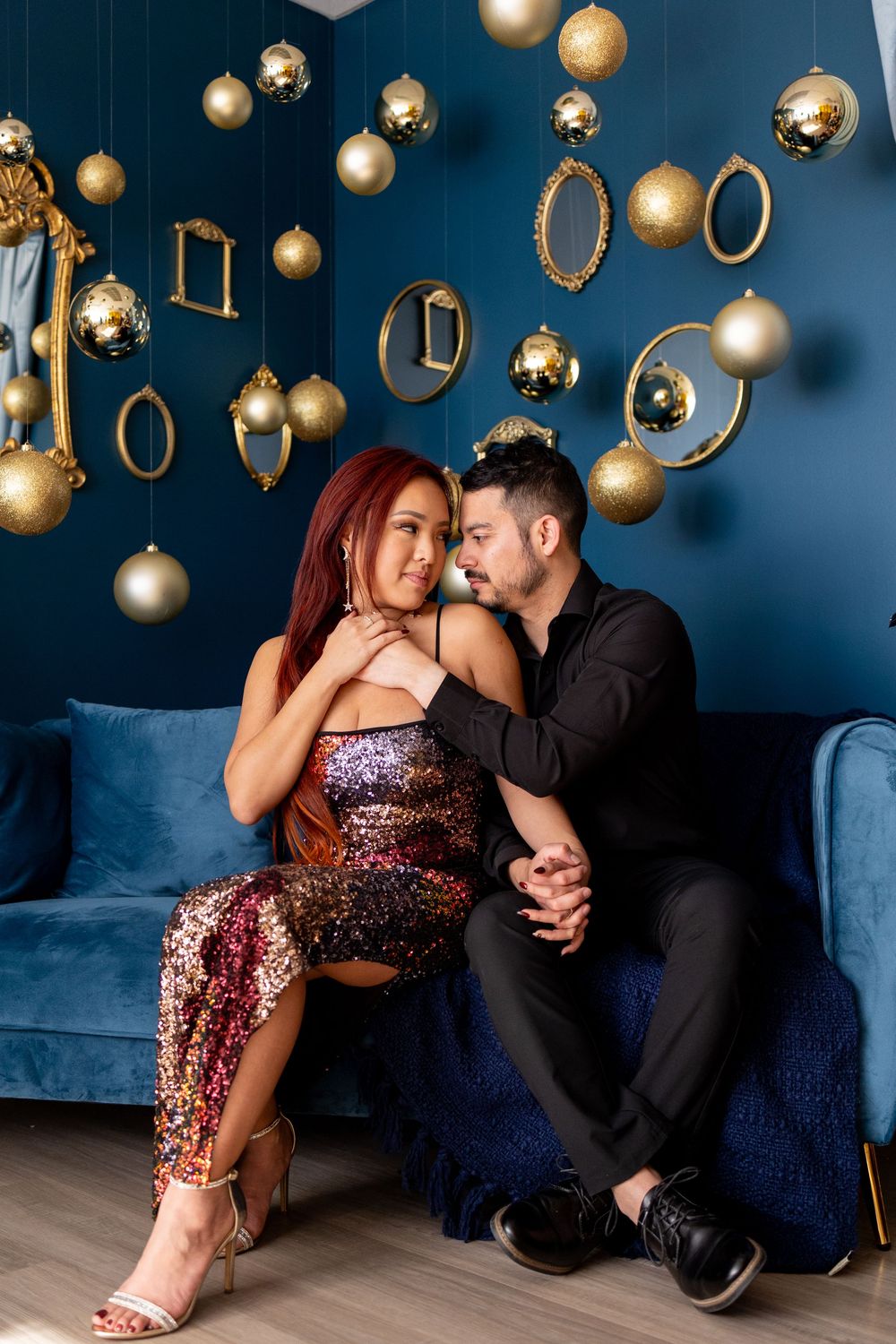 Romantic couple in festive attire sitting on blue velvet sofa with gold Christmas ornaments hanging on teal wall.
