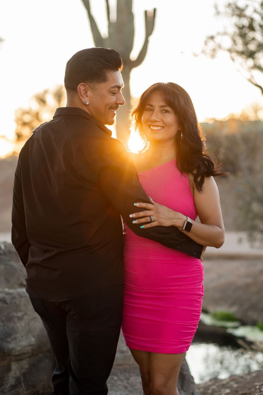 Couple embracing at golden hour sunset in desert landscape with saguaro cactus, woman in pink dress, man in black shirt.