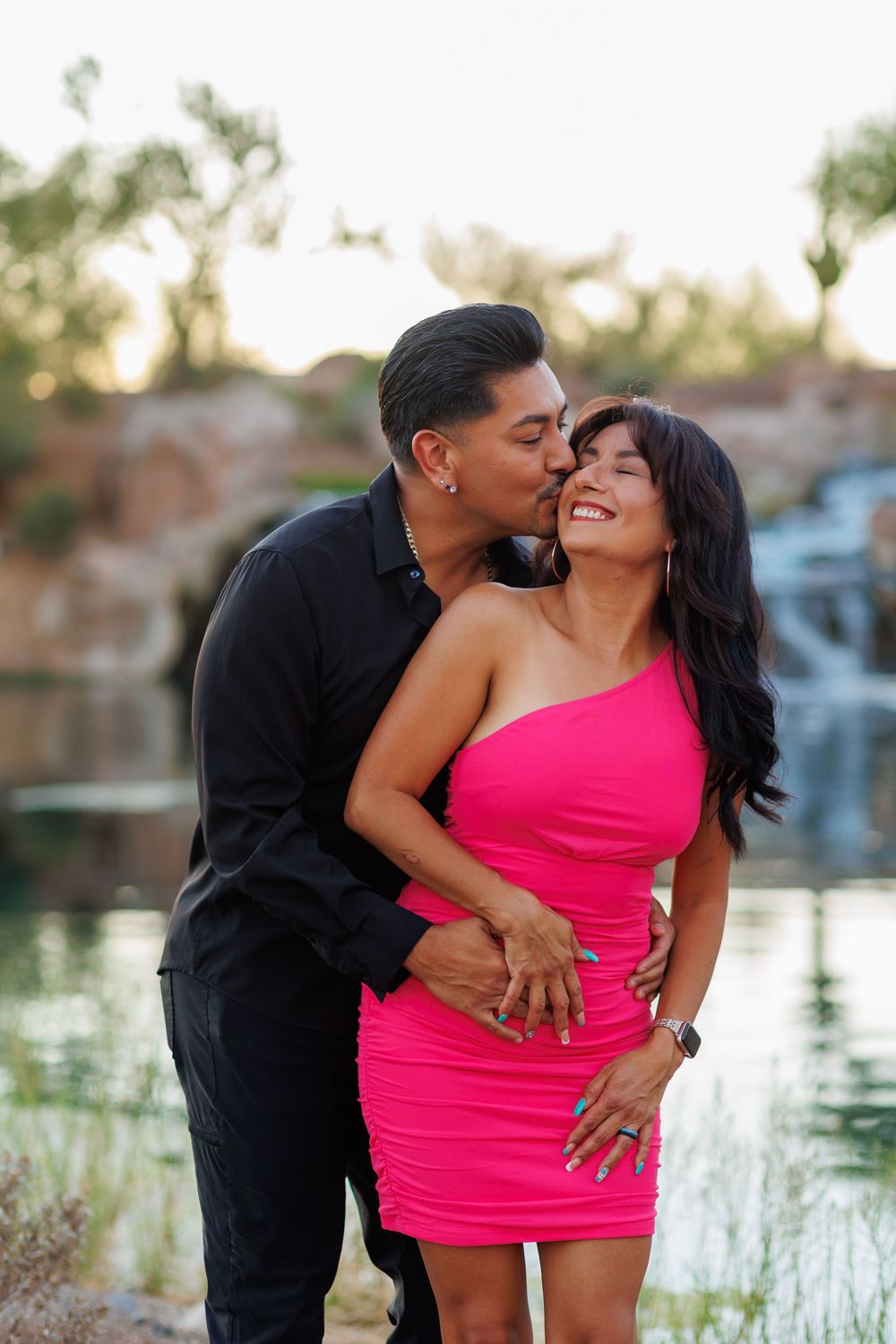 Man in black shirt kissing woman in pink dress on cheek near waterfall, both smiling joyfully outdoors.