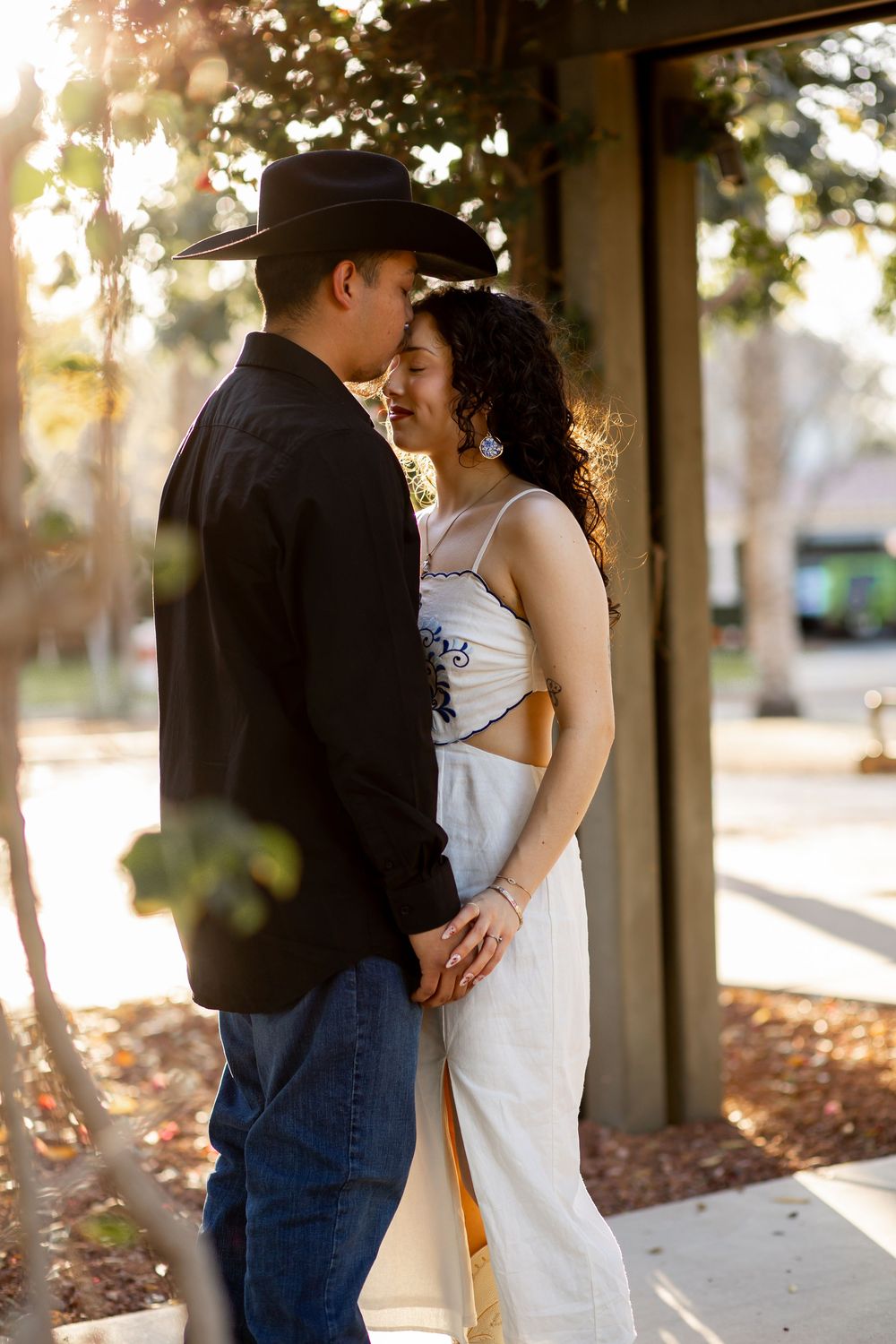 Couple sharing a romantic moment, man in black cowboy hat and shirt, woman in white dress, holding hands outdoors.