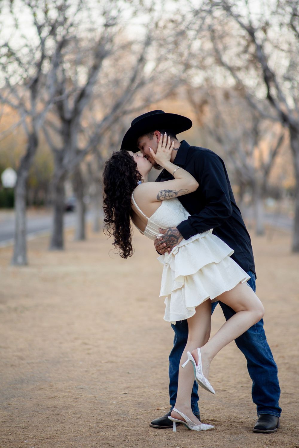 Couple sharing a passionate dip kiss outdoors, man in black cowboy hat, woman in white ruffled mini dress and heels.