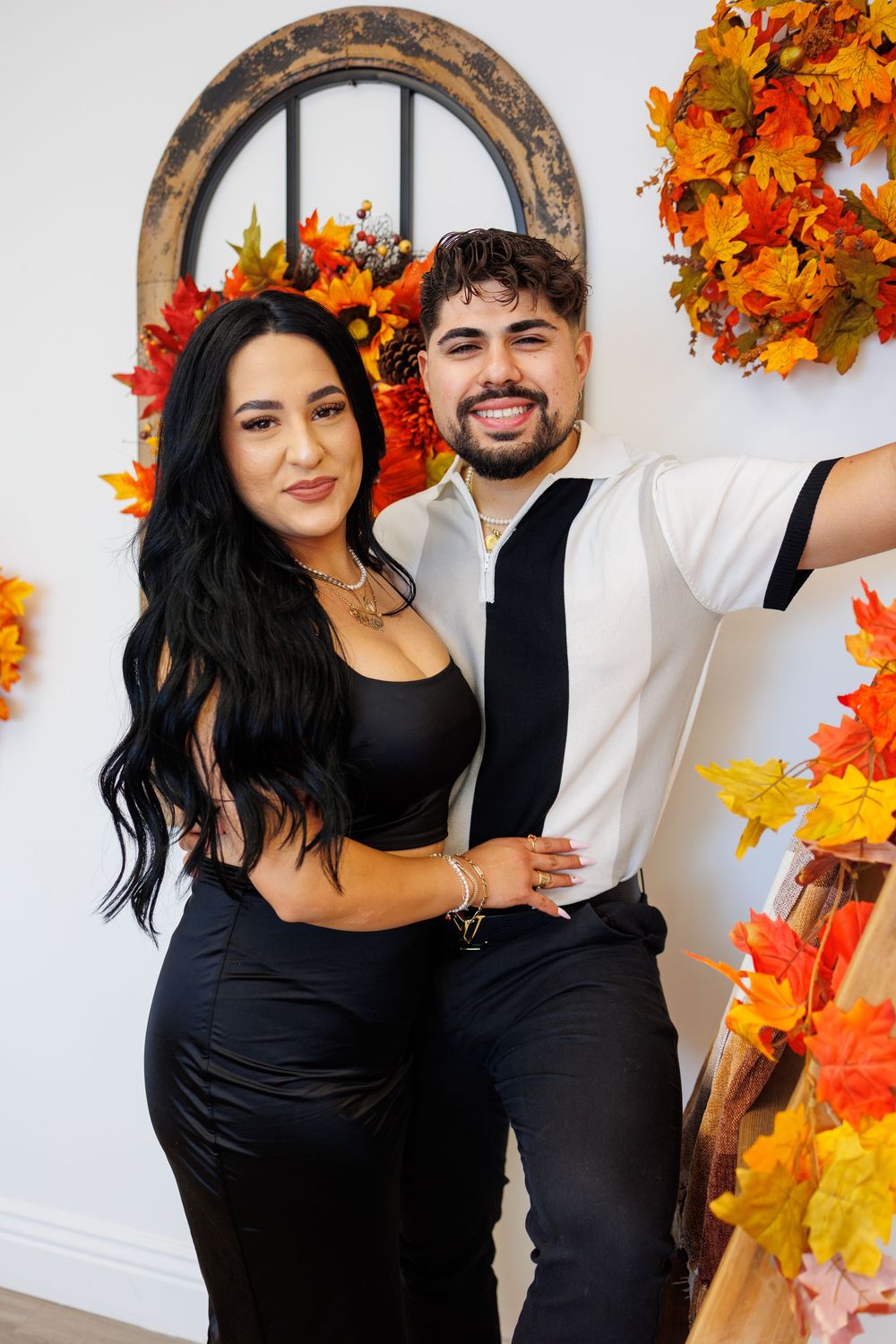 Smiling couple in black outfits posing together in fall-decorated studio setting.