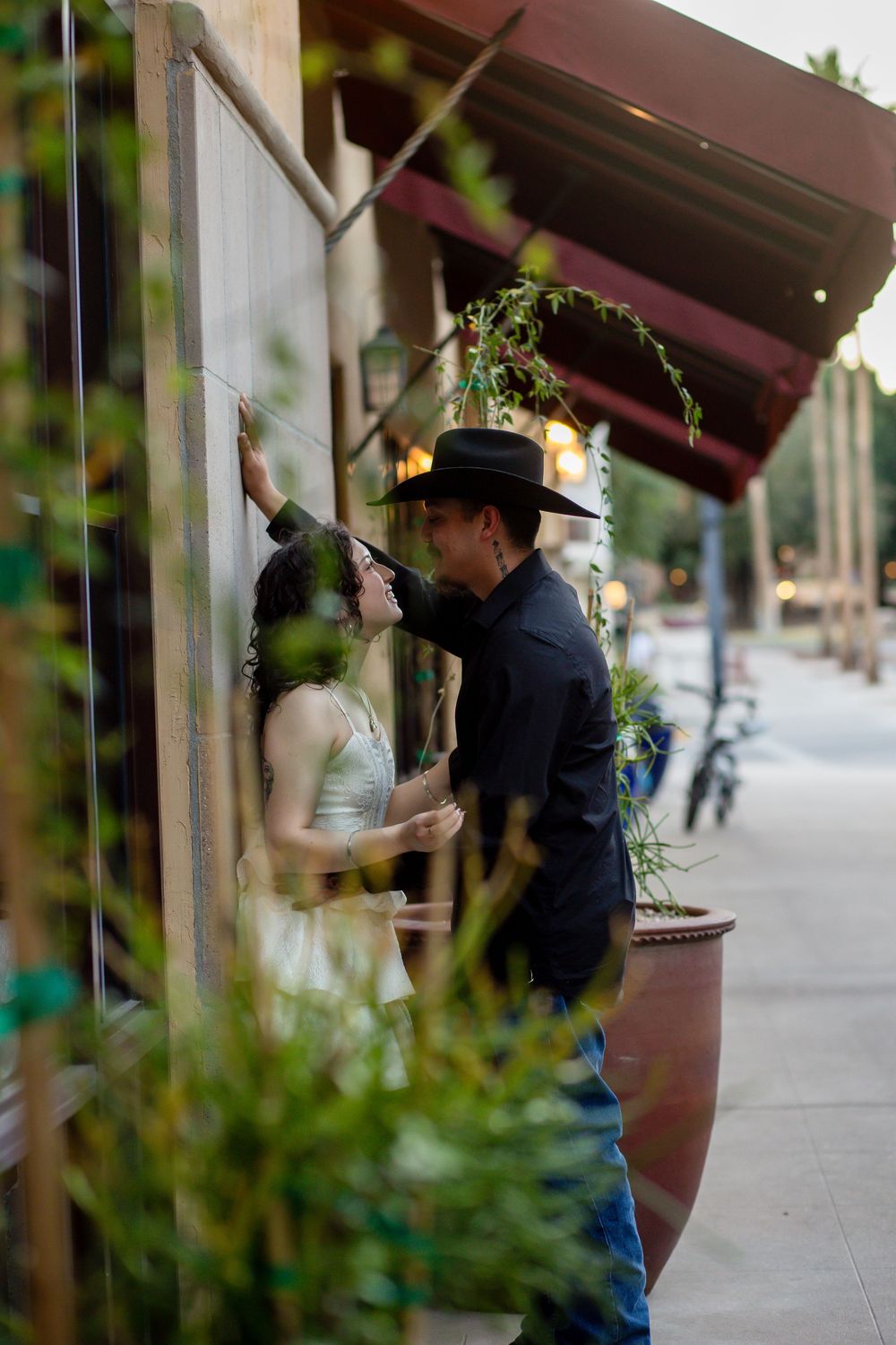 A couple shares a romantic moment outside a restaurant, man in black cowboy hat and jacket leaning toward woman against wall.