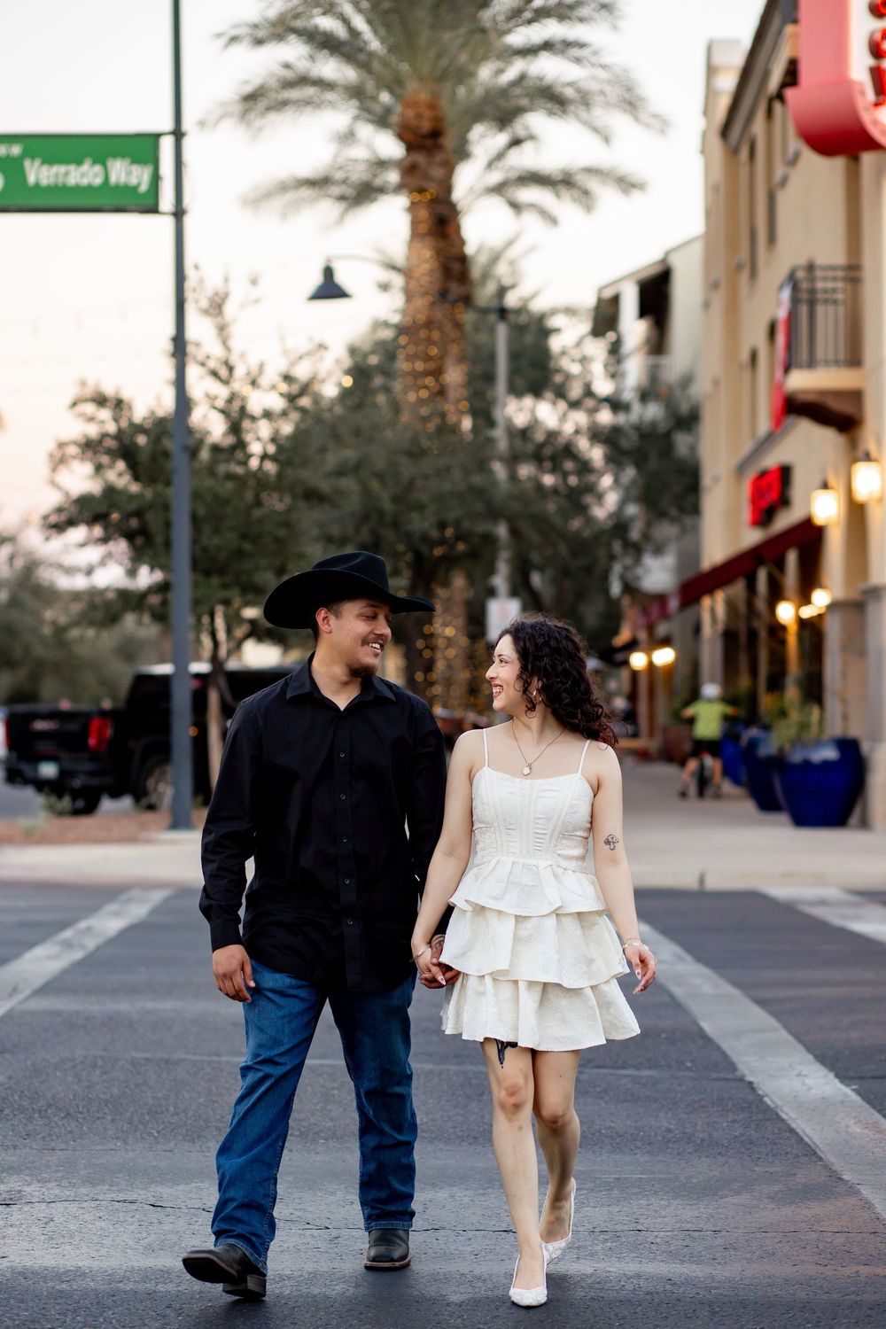 Couple walking hand-in-hand on a street, man in black cowboy hat and shirt, woman in white tiered dress, palm tree background.
