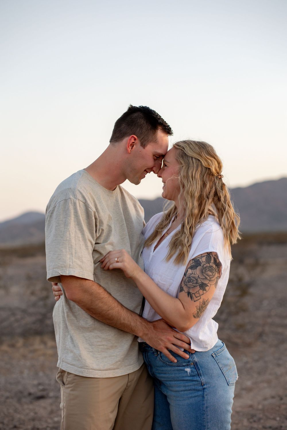 A couple shares a romantic forehead touch in a desert landscape at golden hour, woman with floral arm tattoo in white top.