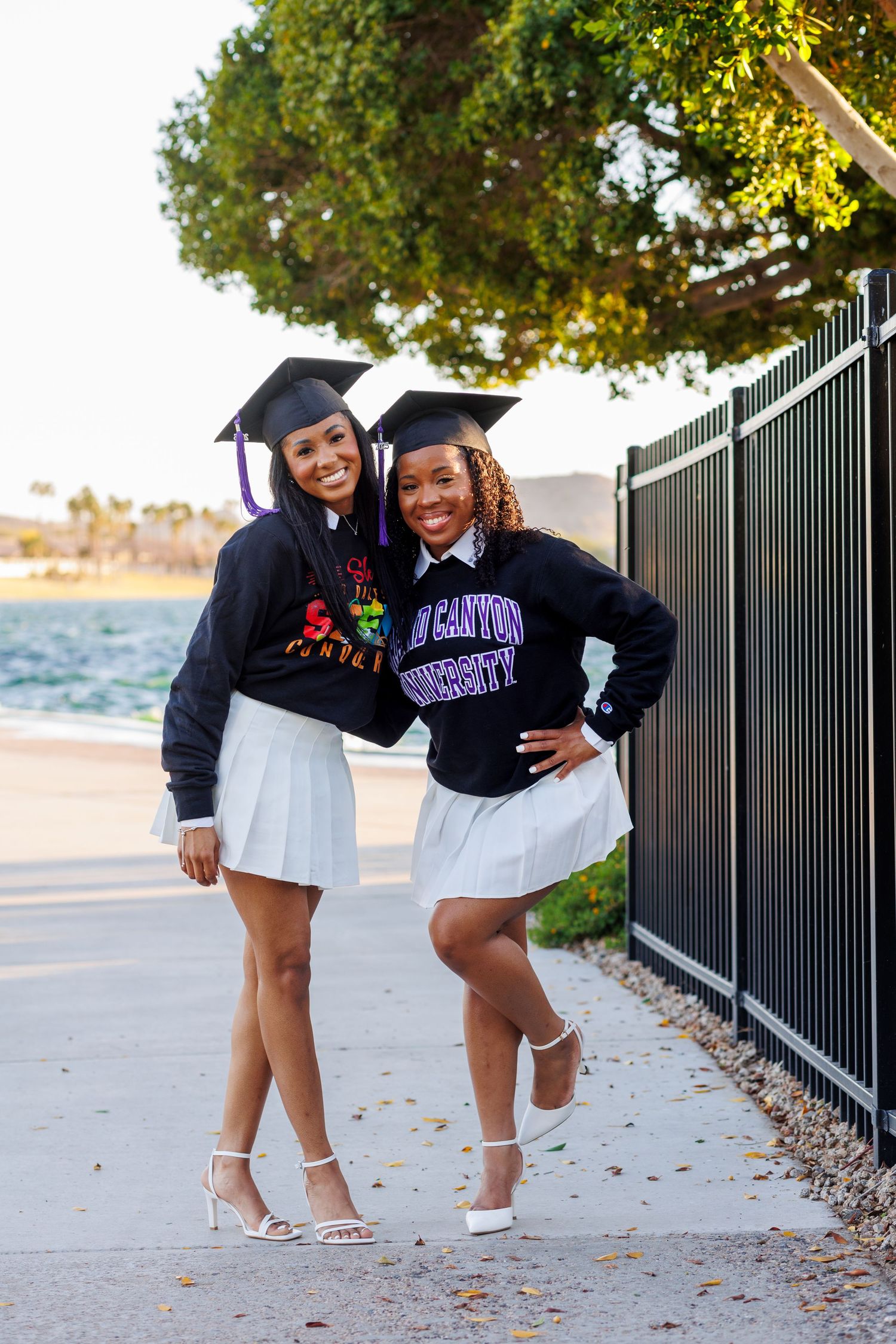 Two graduates in caps and sweatshirts pose proudly in white skirts by the waterfront.