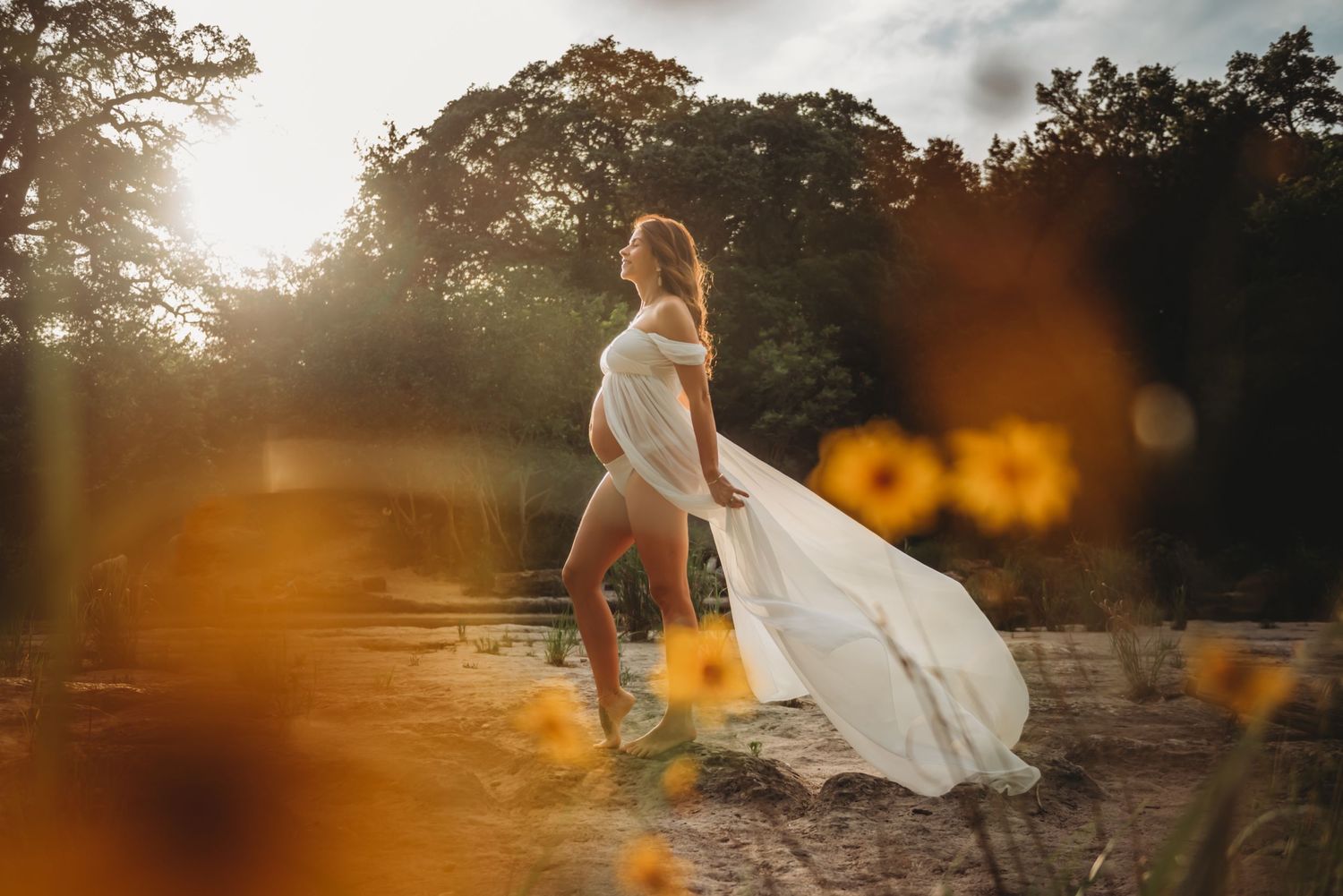 A bride in a flowing white dress stands among yellow flowers at sunset, with dreamy bokeh effects in the foreground.