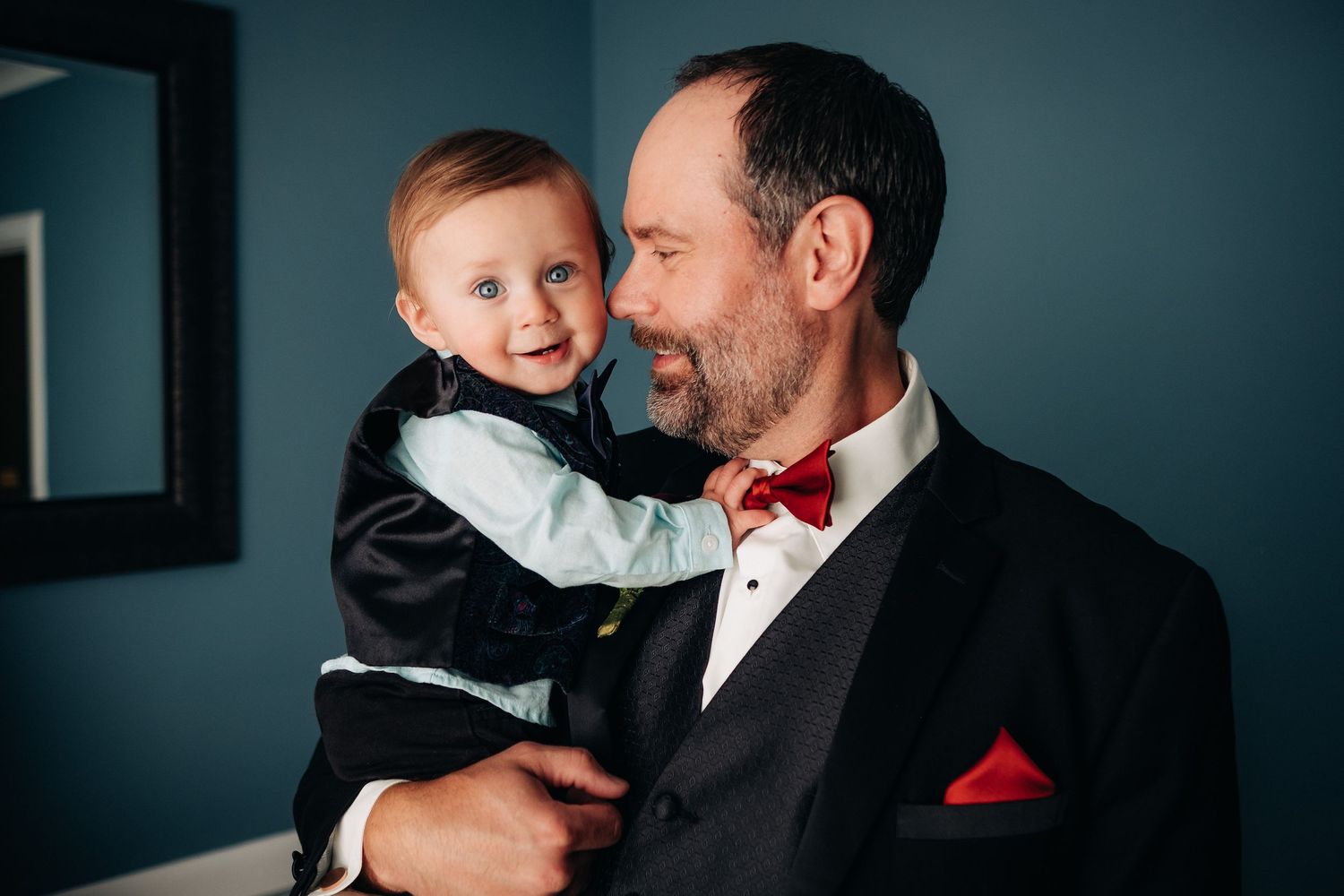 Tender moment between two people in formal attire with bow tie against blue wall.