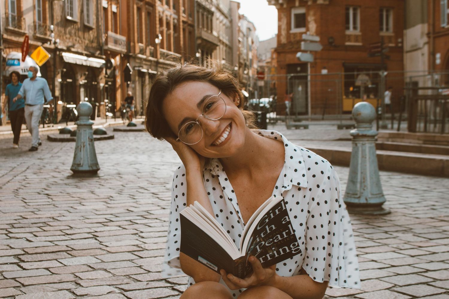 Person reading a book on cobblestone street with classic European architecture in background.