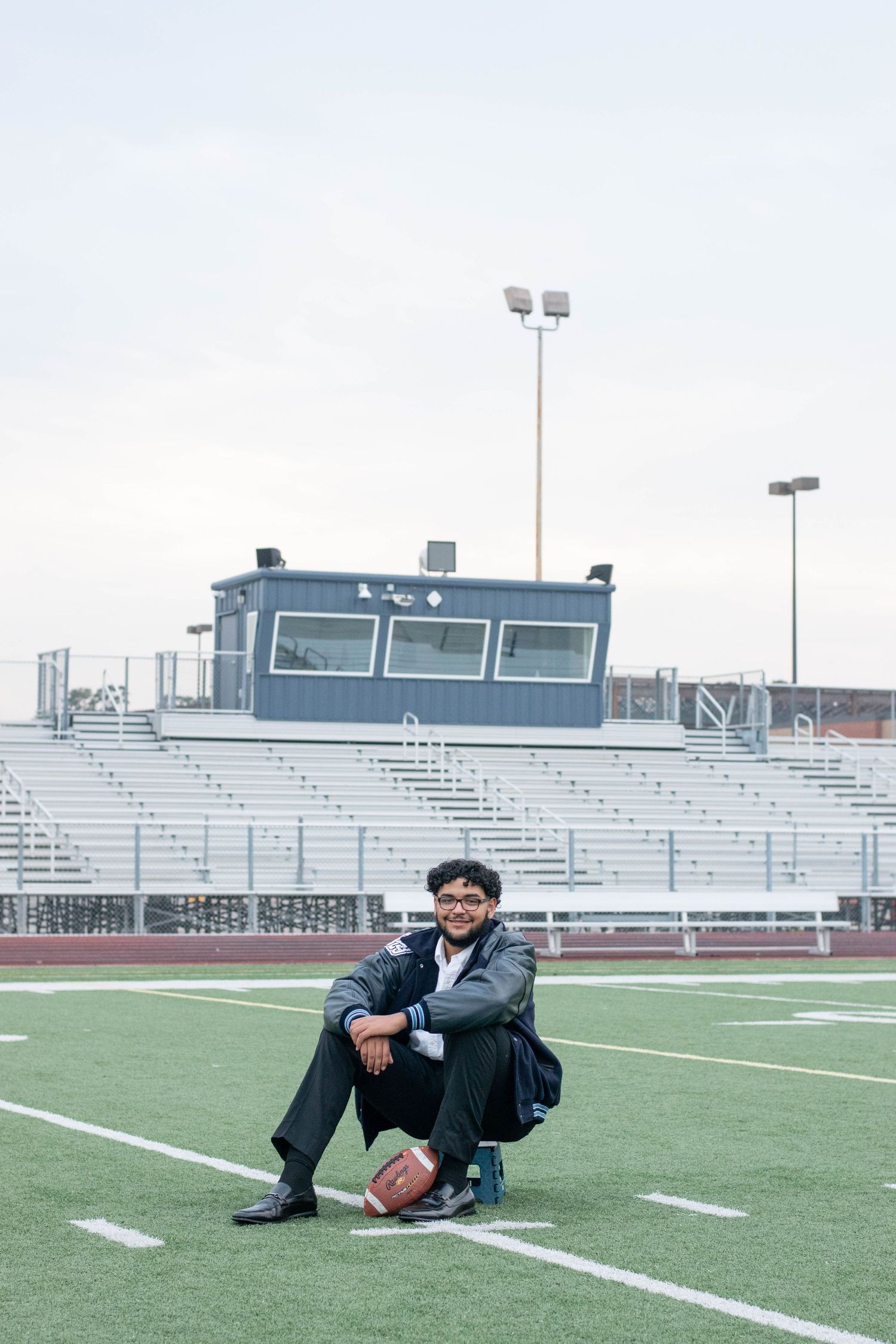 Athletic figure poses with football on field with stadium bleachers in background.