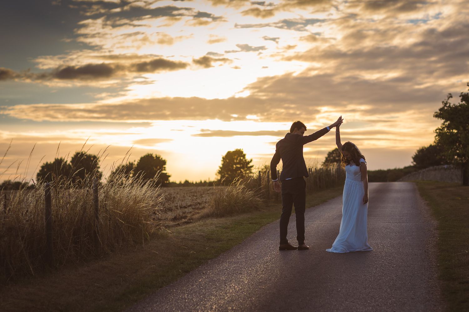 A couple stands on a country road during golden hour sunset with dramatic clouds.
