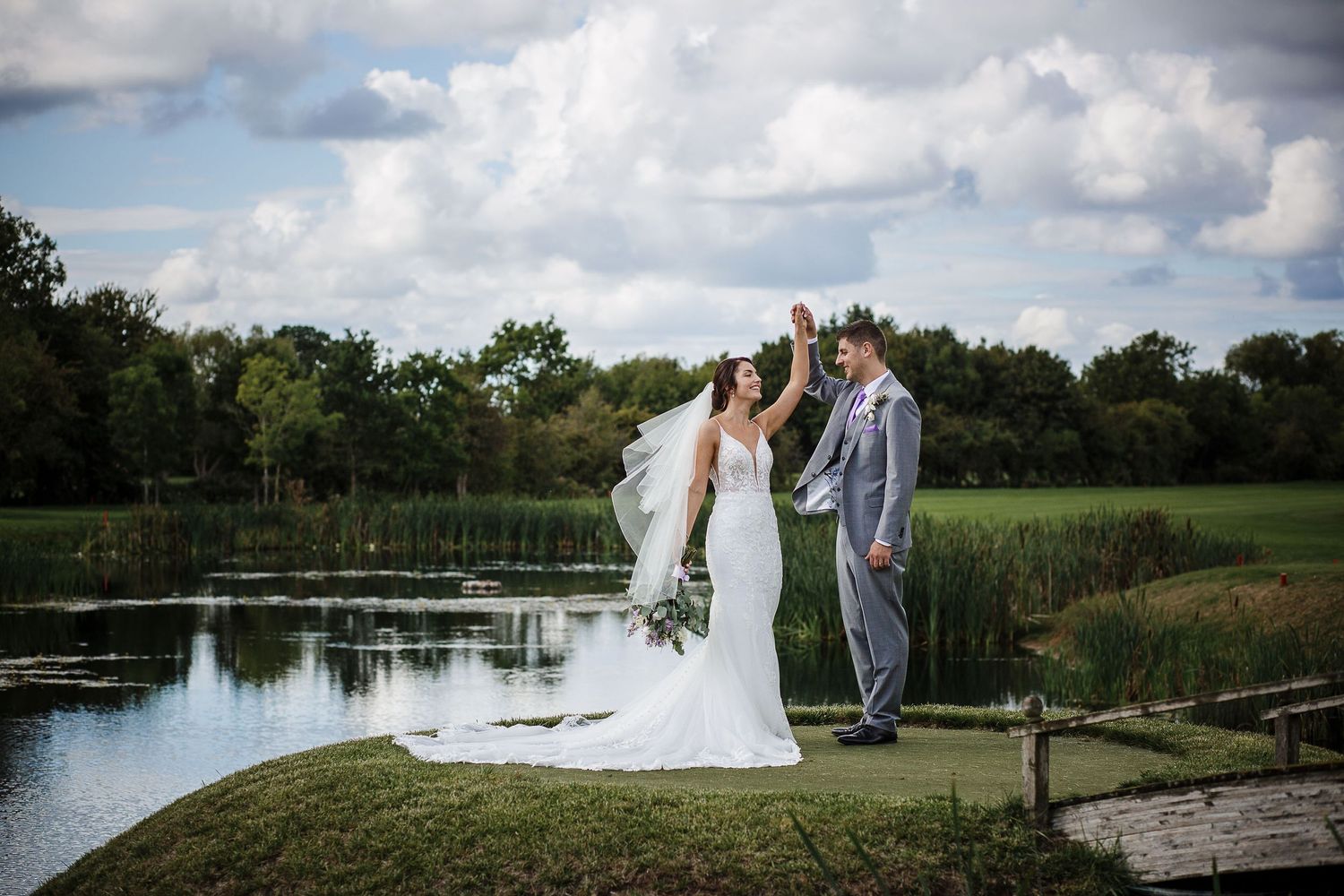 Newlyweds share intimate moment by scenic pond with lush greenery on wedding day.
