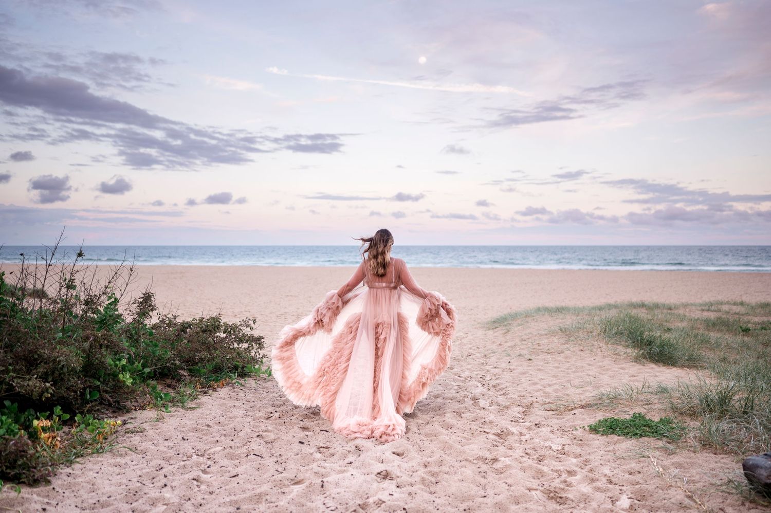 Maternity photo on a beach at sunset with flowing pink dress and cloudy sky.