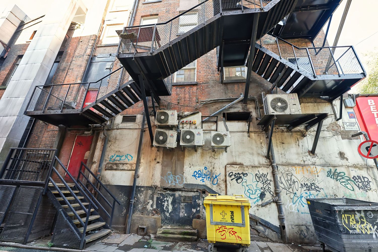 Graffiti-covered urban alleyway with fire escapes and metal stairs on brick buildings.