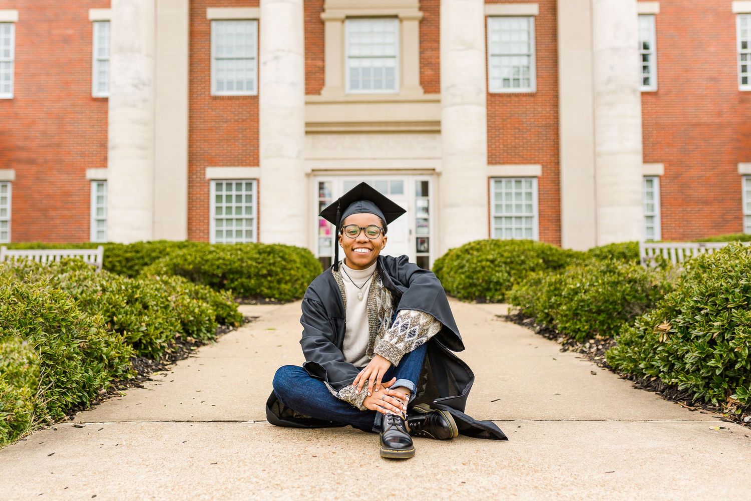 Graduate in cap and gown sits casually in front of a brick academic building.
