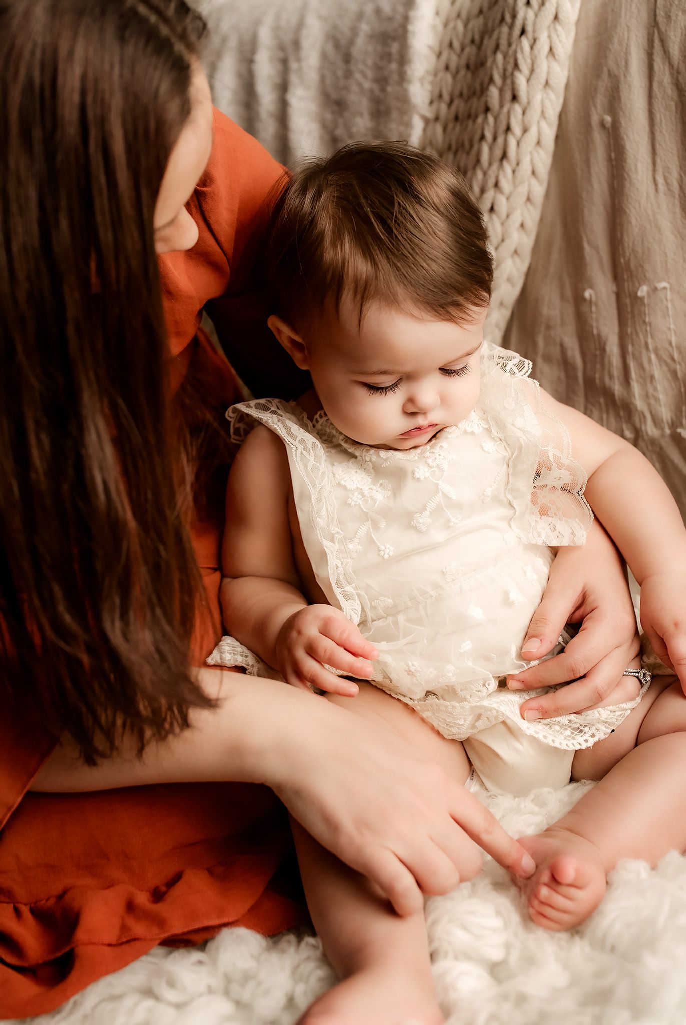 Mother in red dress tenderly holds baby girl in white lace dress on fluffy white blanket.