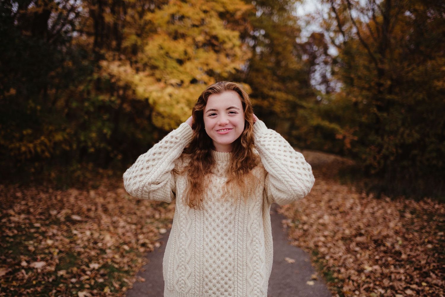 A person in a cream sweater smiles joyfully among autumn leaves.