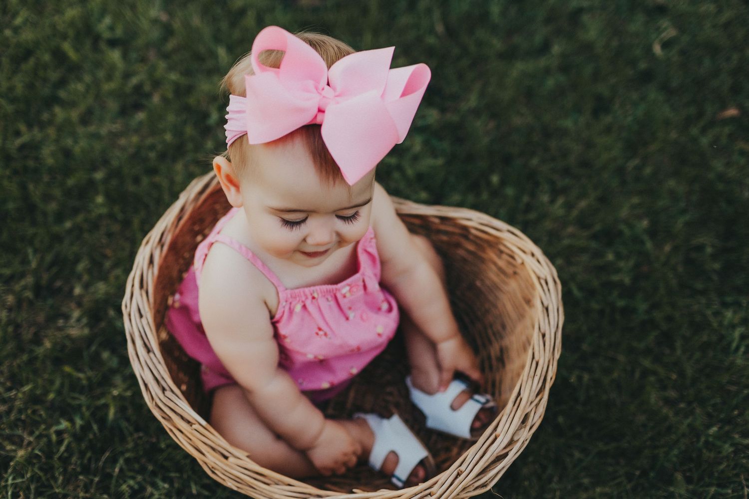 A baby wearing pink sits in a woven basket on green grass.