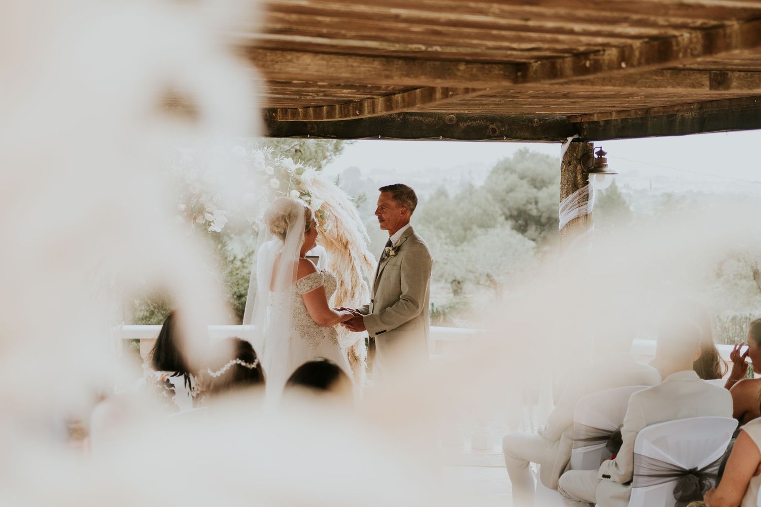 Wedding ceremony takes place under a wooden pergola with guests seated in white chairs.