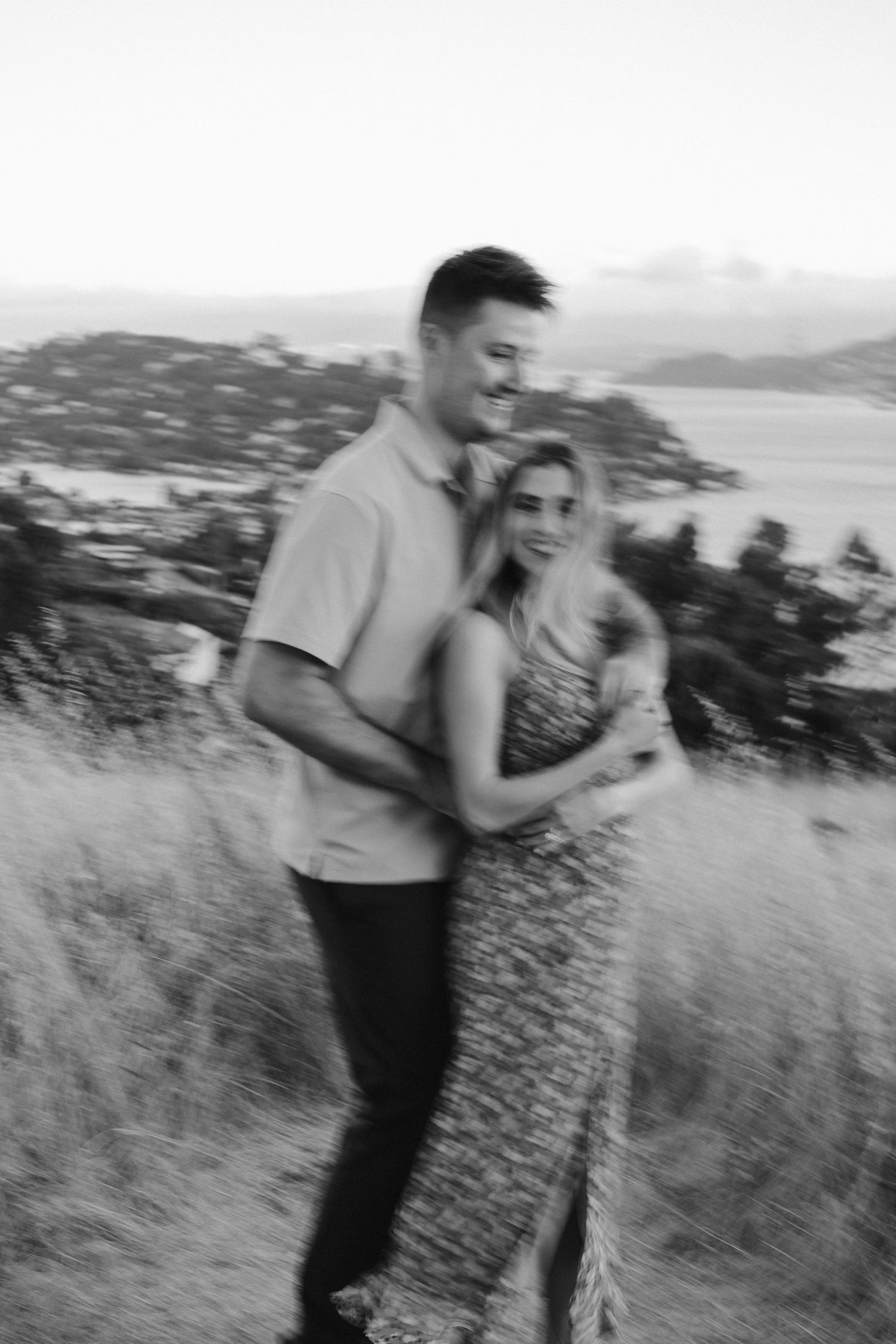 A romantic black and white portrait of a couple embracing on a hilltop with a scenic vista in the background.