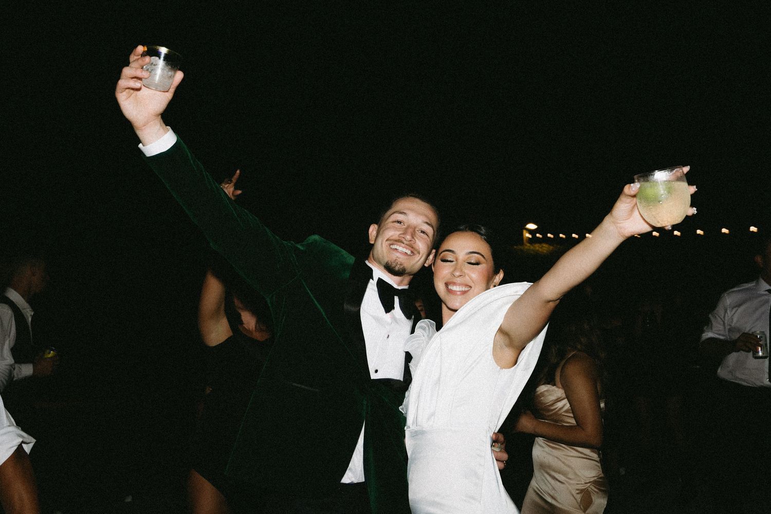Bride and groom raise cocktails and celebrate together at an outdoor evening wedding reception.