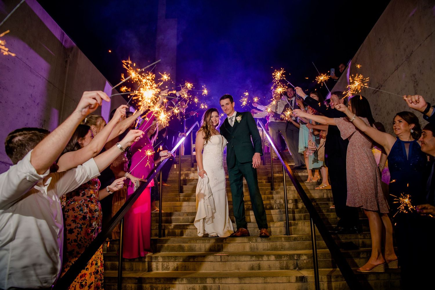 Newlyweds walk down stone steps through a sparkler tunnel held by wedding guests at night.
