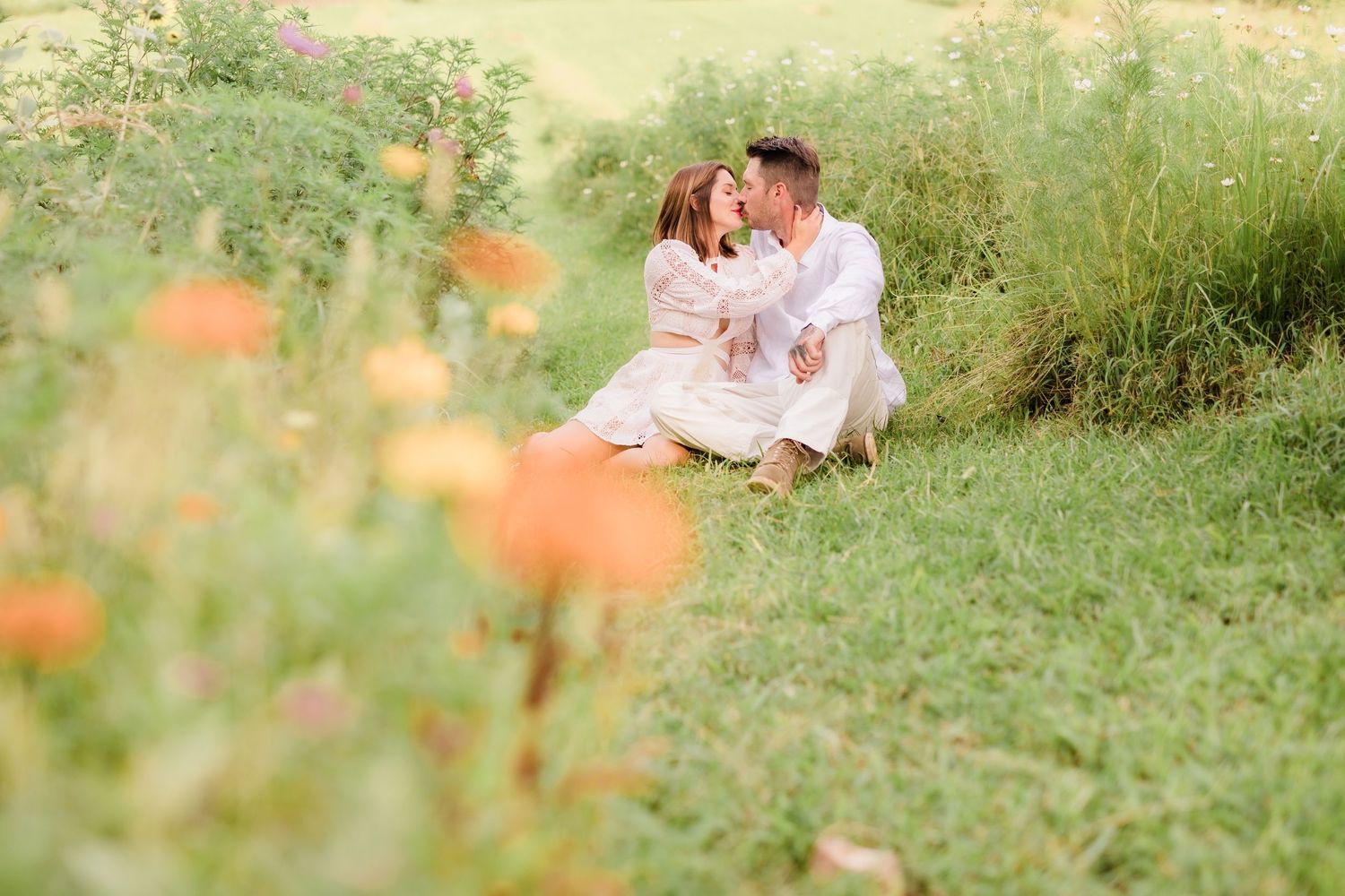 Romantic couple sharing a kiss while sitting in a lush green meadow surrounded by colorful wildflowers.