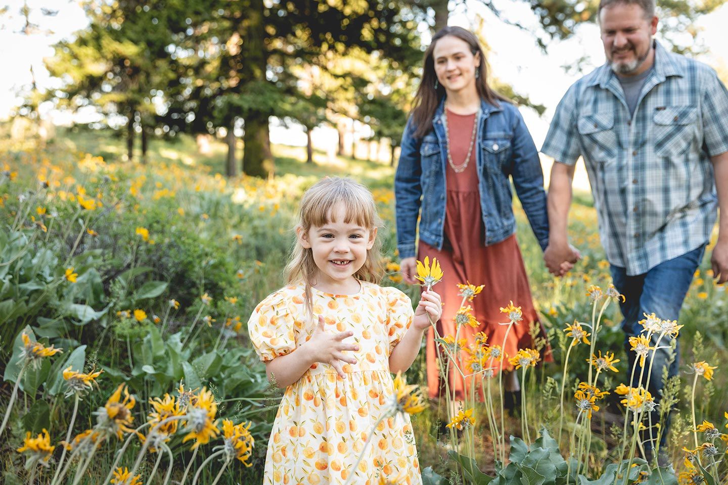 young girls smiling while standing in mountain wildflowers and parents walking up behind her