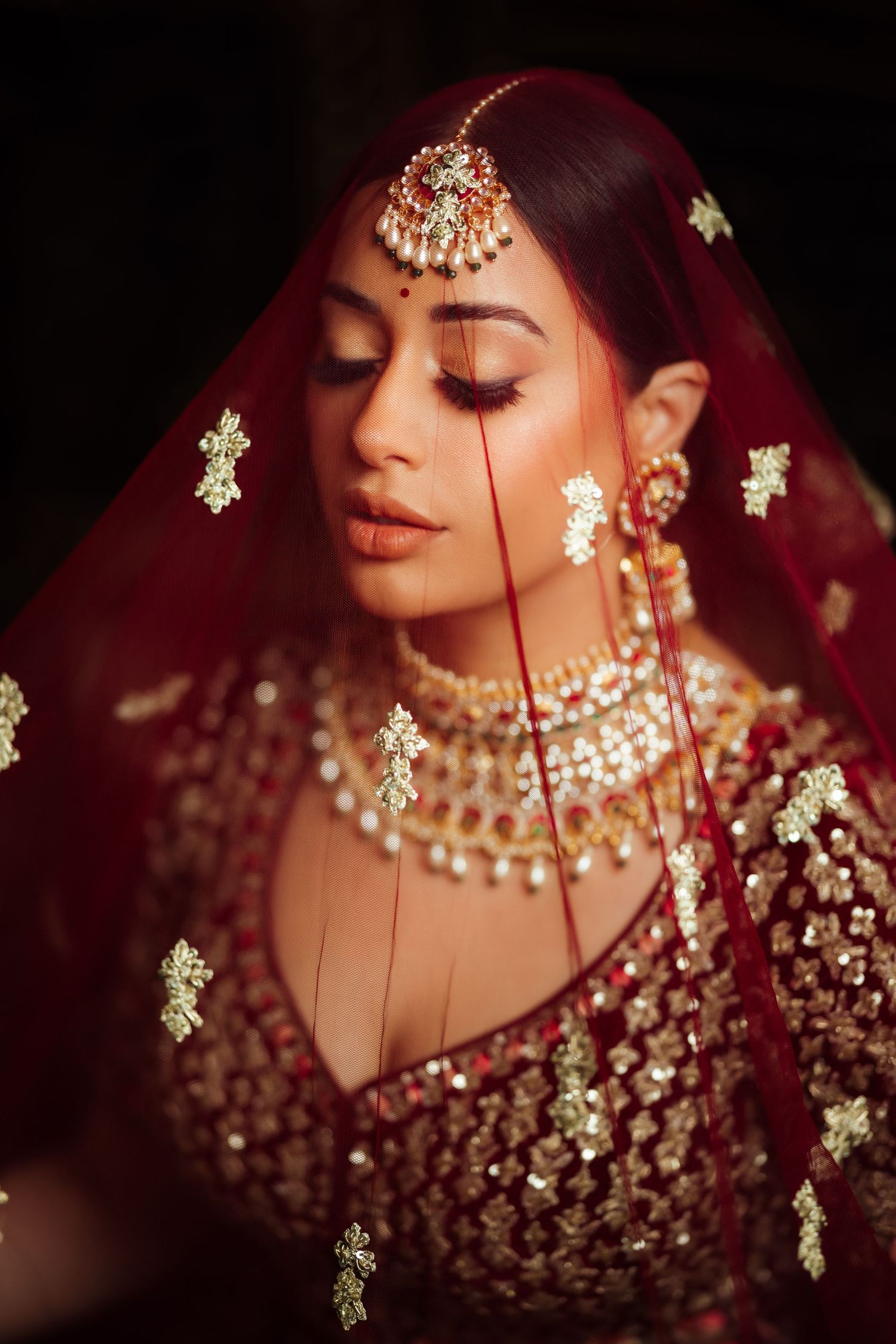 Close-up portrait of bride in red veil with ornate gold jewelry and traditional Indian bridal makeup.