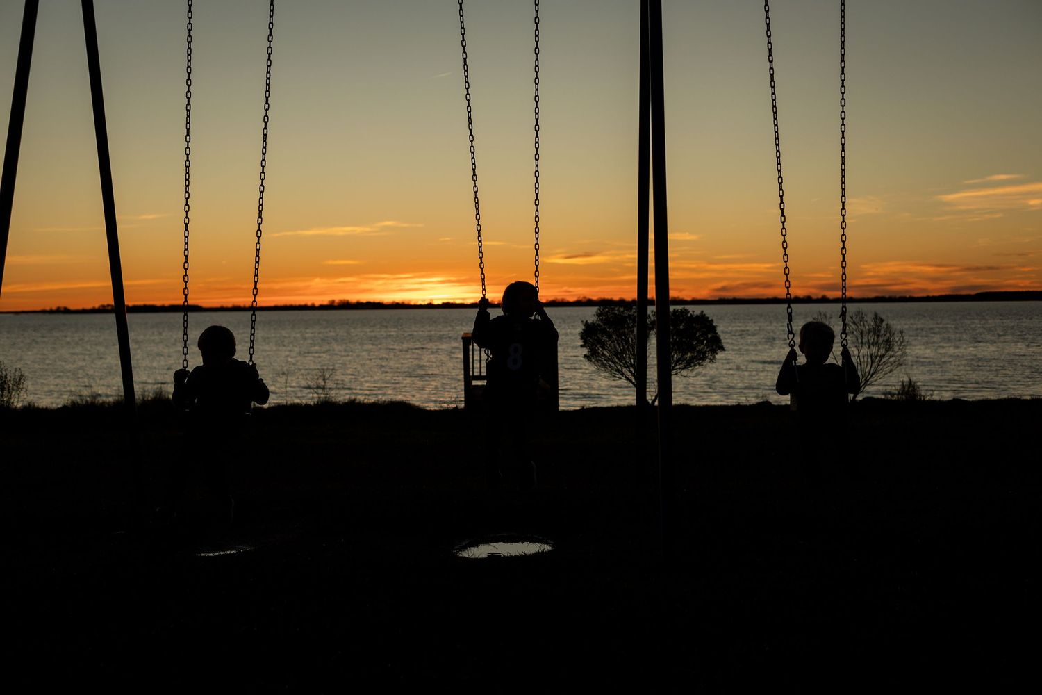 Silhouettes of people on swings against a vibrant orange sunset over water.