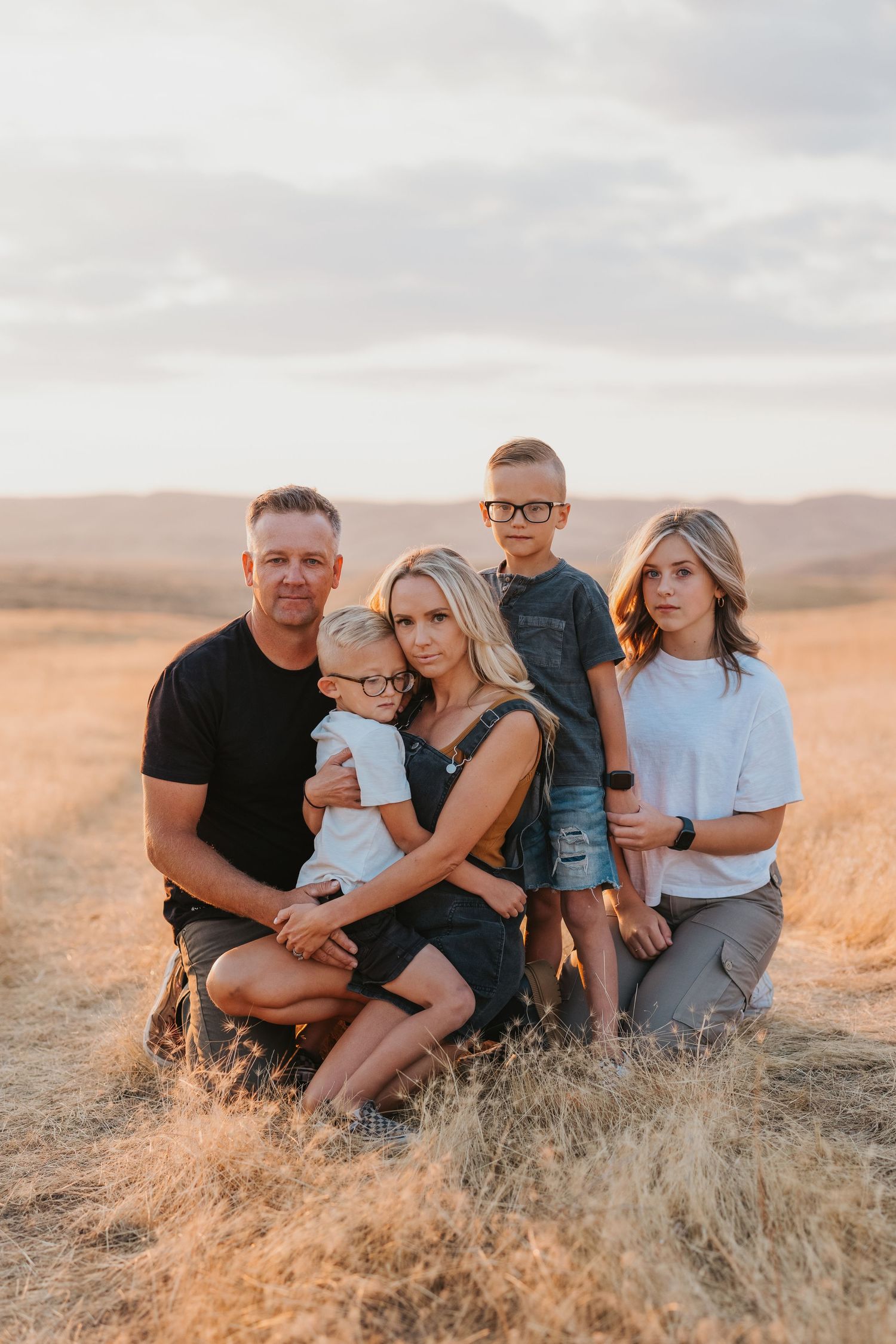 A family of five poses together in casual attire against a backdrop of golden fields at sunset.