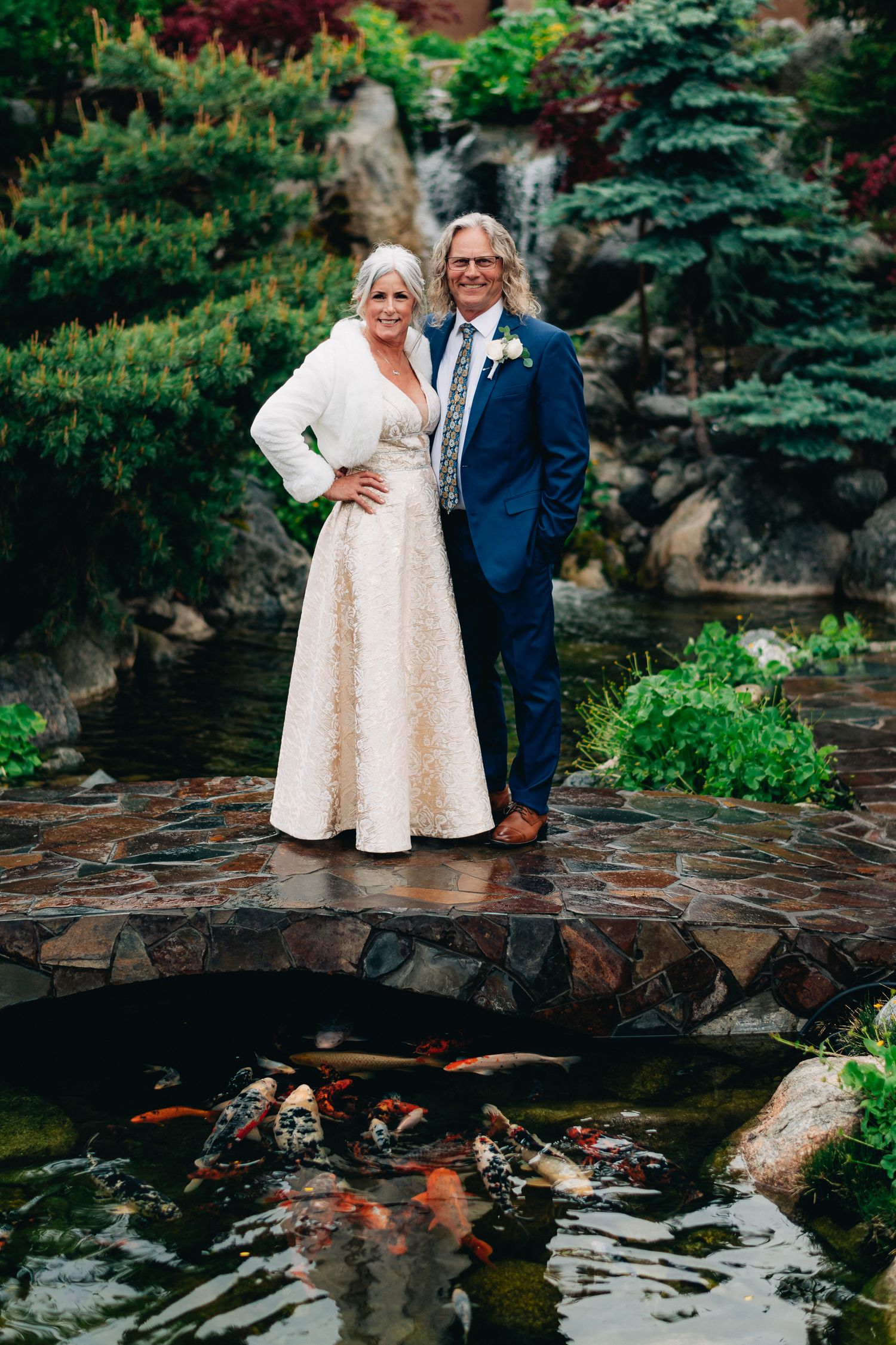 Couple in formal attire posing on stone bridge over koi pond in garden setting.