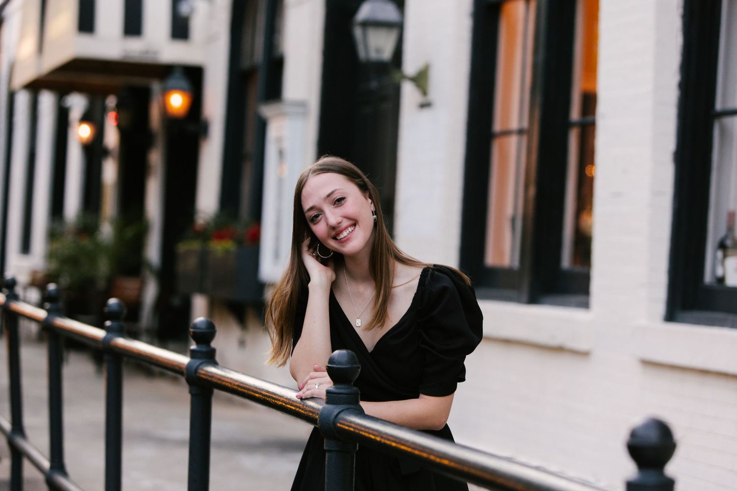 A woman in a black dress poses along an ornate railing near historic buildings at dusk.