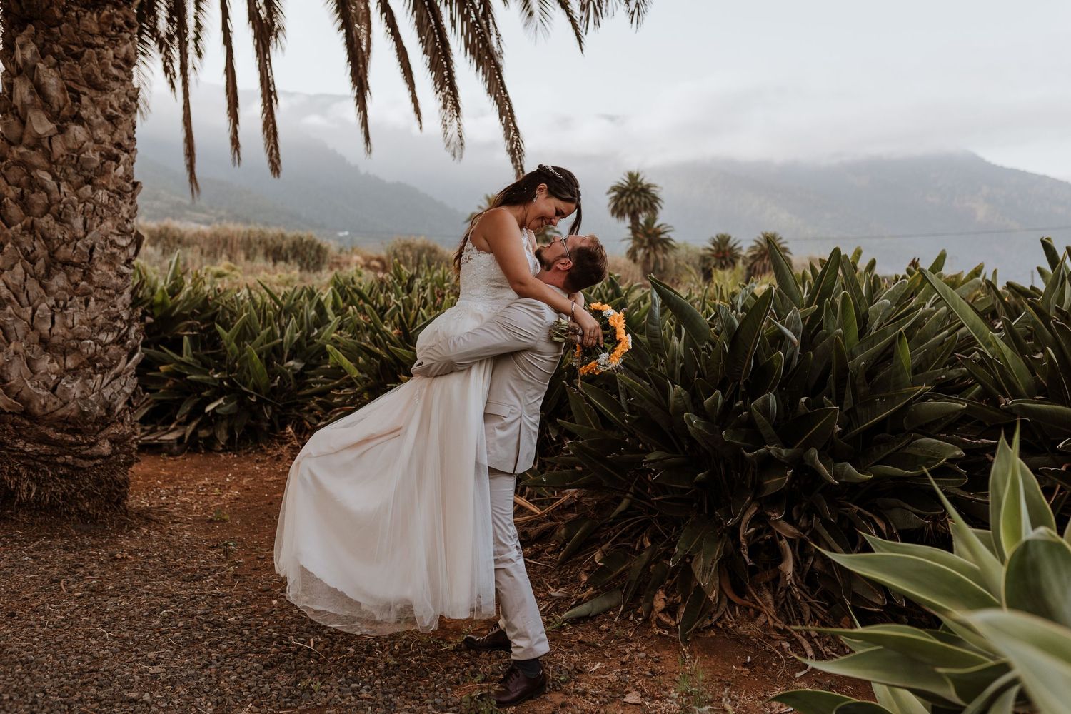Wedding couple embracing at sunset with palm trees and mountains in the background.