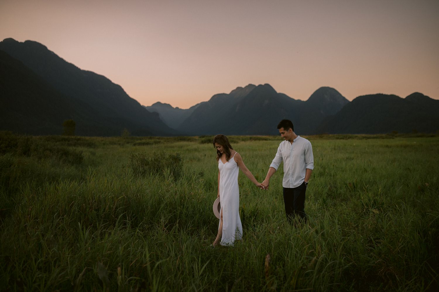 Couple walking through a lush green meadow with mountains in the background at sunset.
