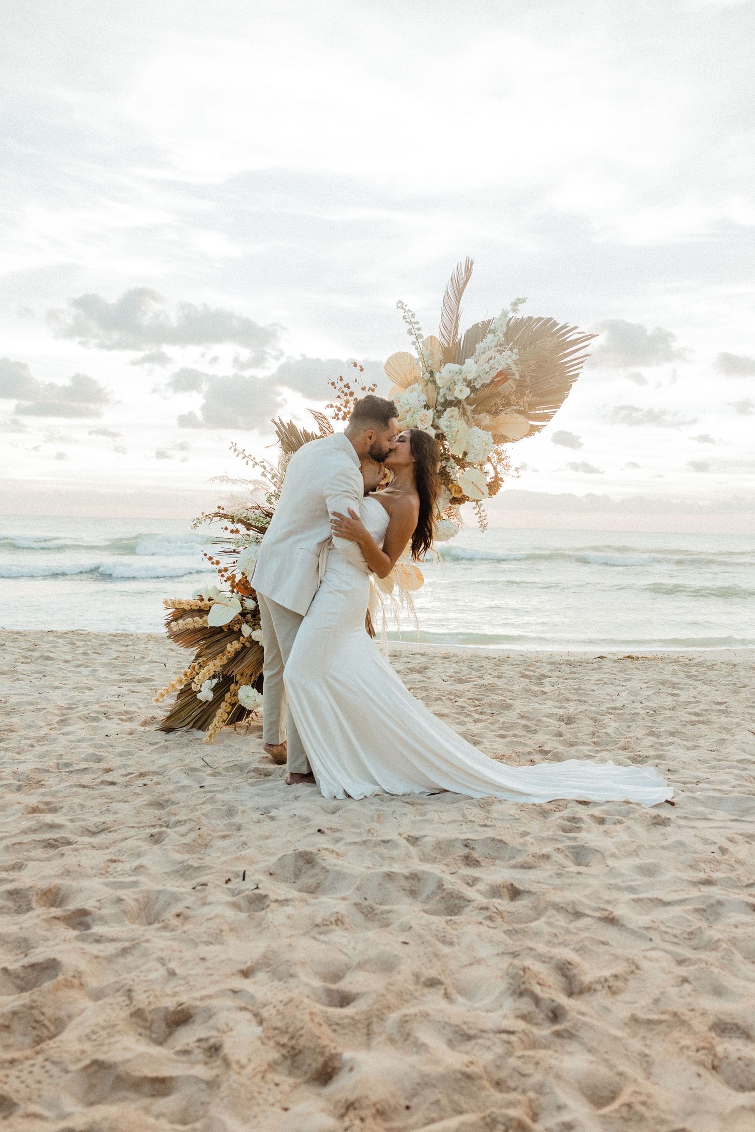 Bride and groom share a romantic dip kiss on sandy beach before a boho floral arch at sunset.