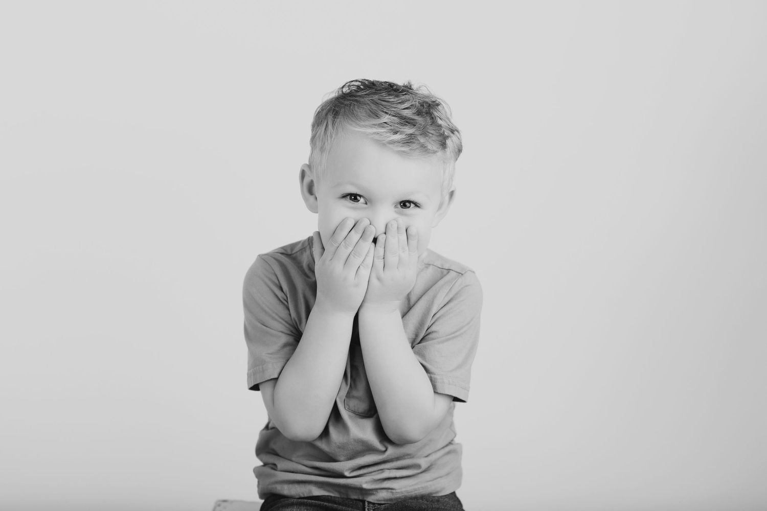 Young subject in gray t-shirt covers face with hands in black and white portrait photo.