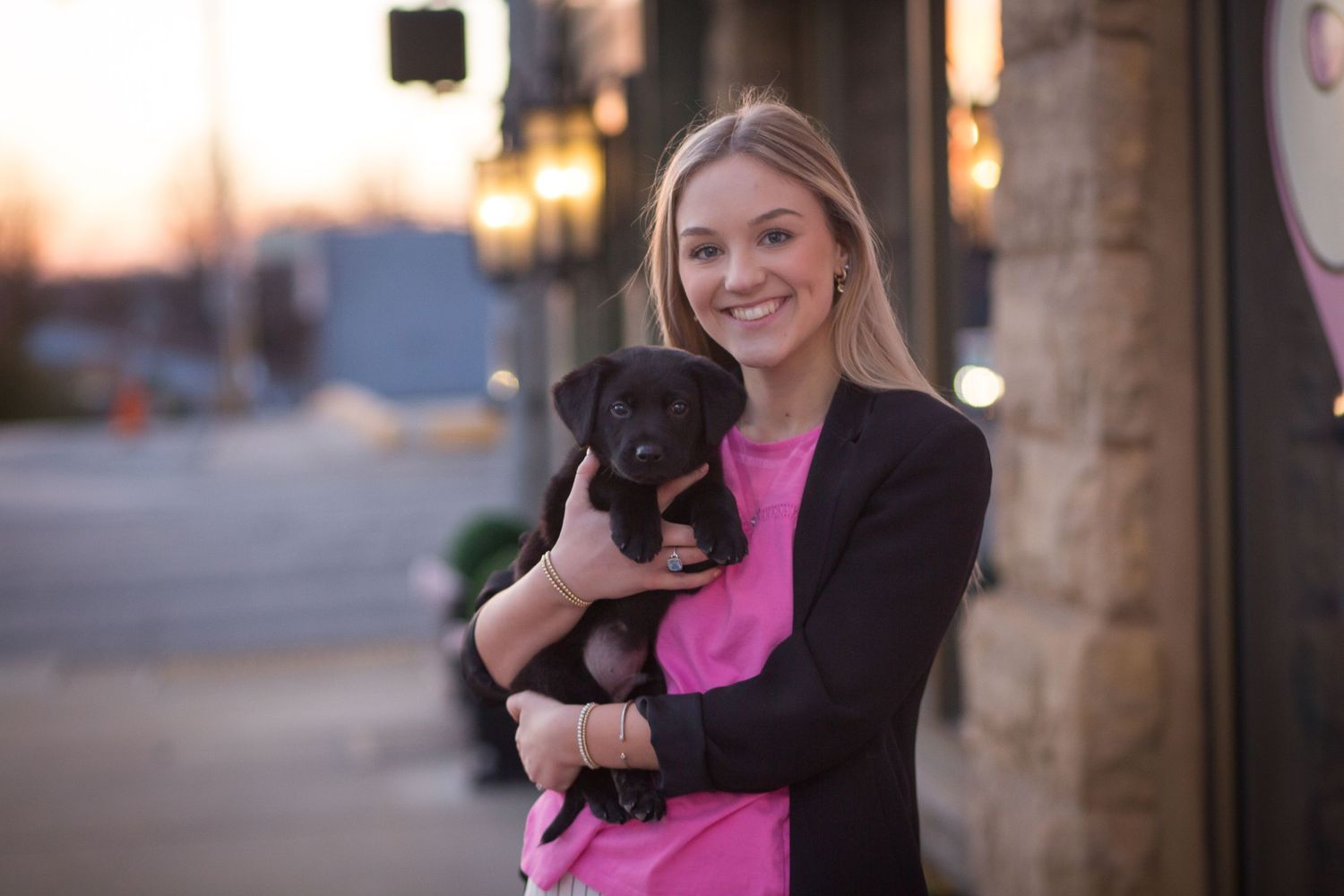 Smiling young woman in pink top and blazer holding a small black puppy outdoors at sunset.