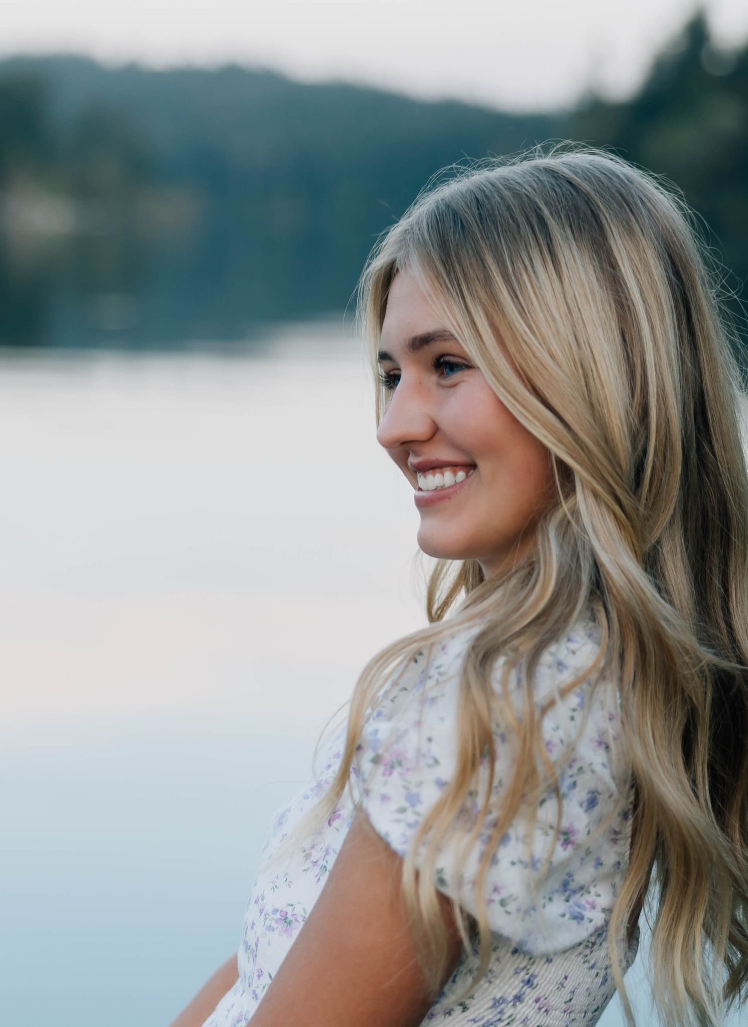Smiling blonde woman with wavy hair near a lake at dusk in floral dress.