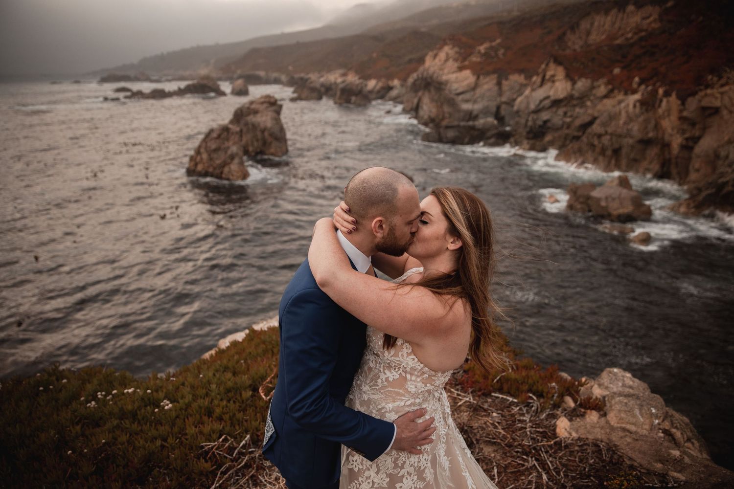 A romantic wedding photo captured along a dramatic rocky coastline at sunset with crashing waves below.