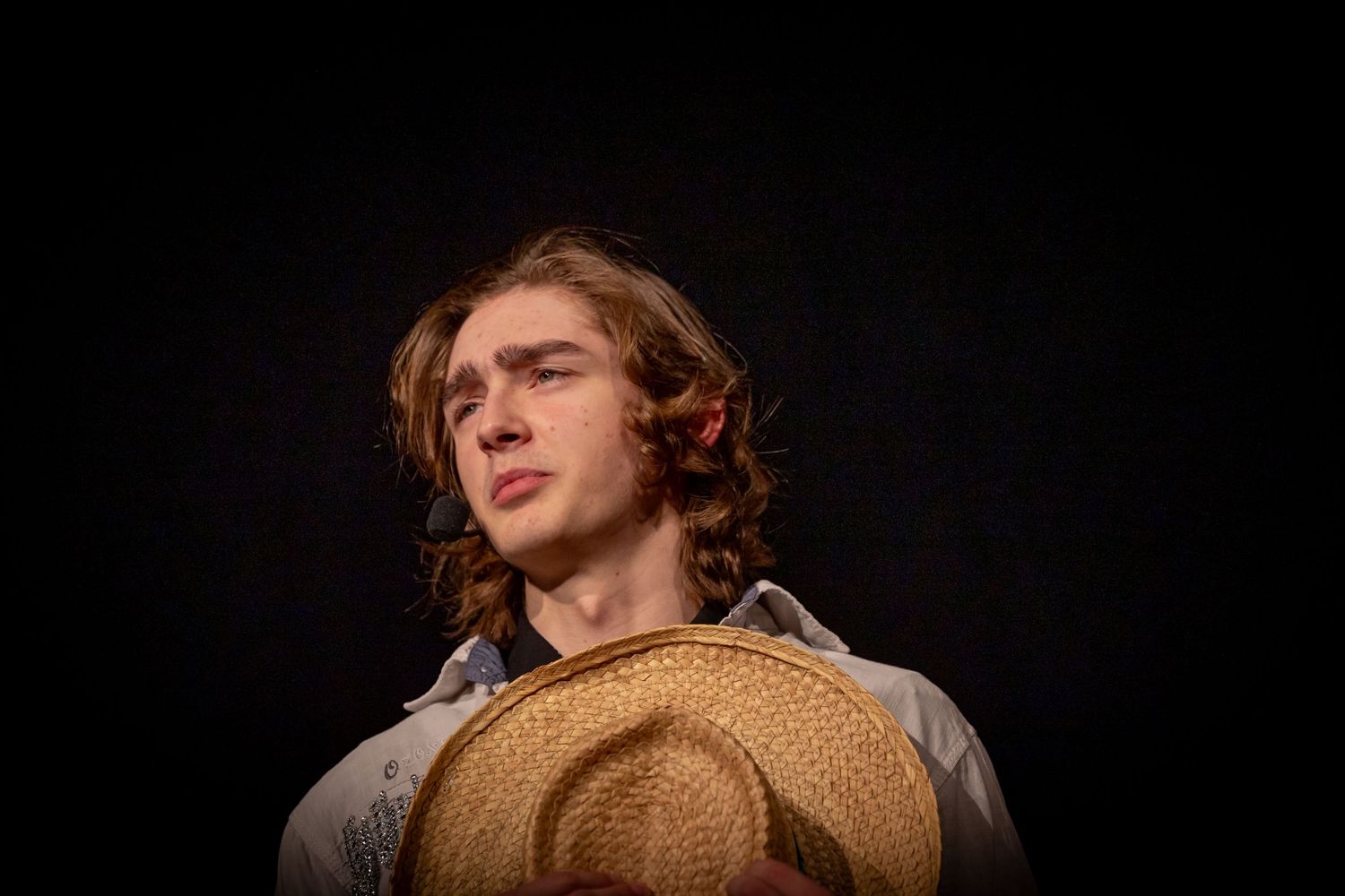 Dramatic portrait of person holding straw hat against black background.