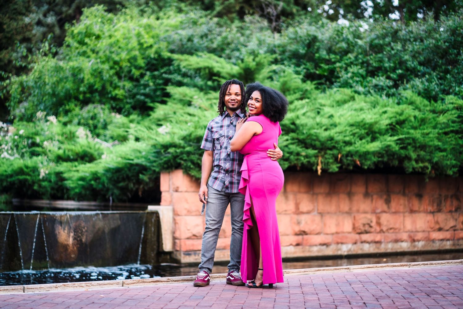 A couple poses together near a brick fountain in a lush garden setting wearing contrasting casual and formal attire.