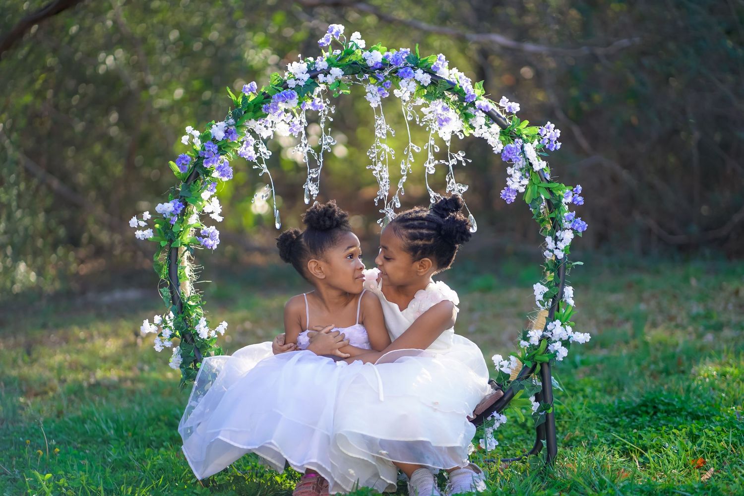 Two children in white dresses share a sweet moment under a floral arch decorated with blue and white flowers.