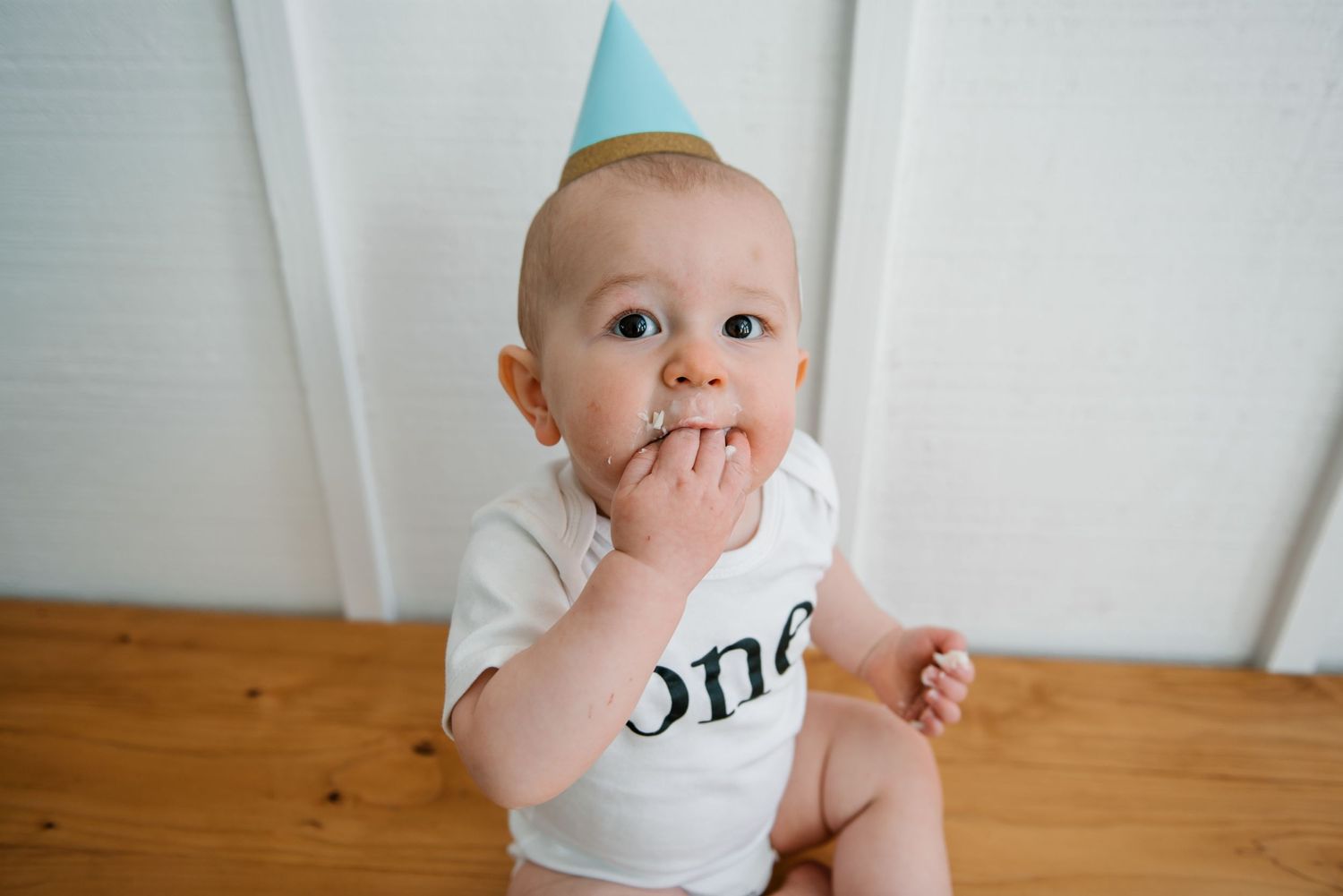 A baby in white clothing sits against a white wall background.
