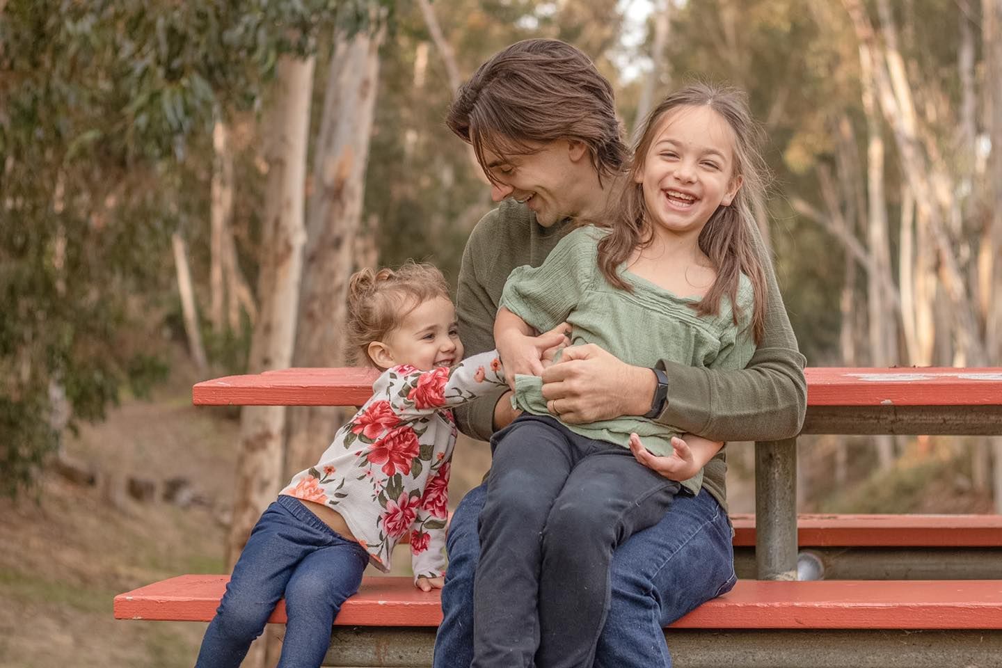 Three people sharing a playful moment on a red bench surrounded by eucalyptus trees.