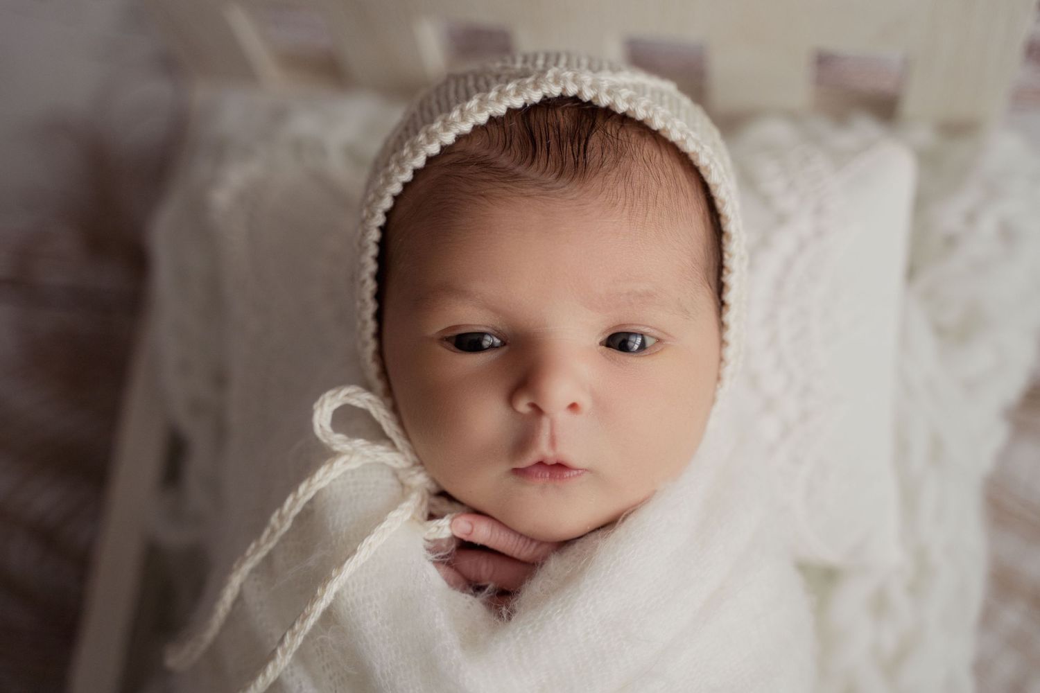 Adorable newborn baby in a white knit bonnet and wrap gazes at the camera with big blue eyes.