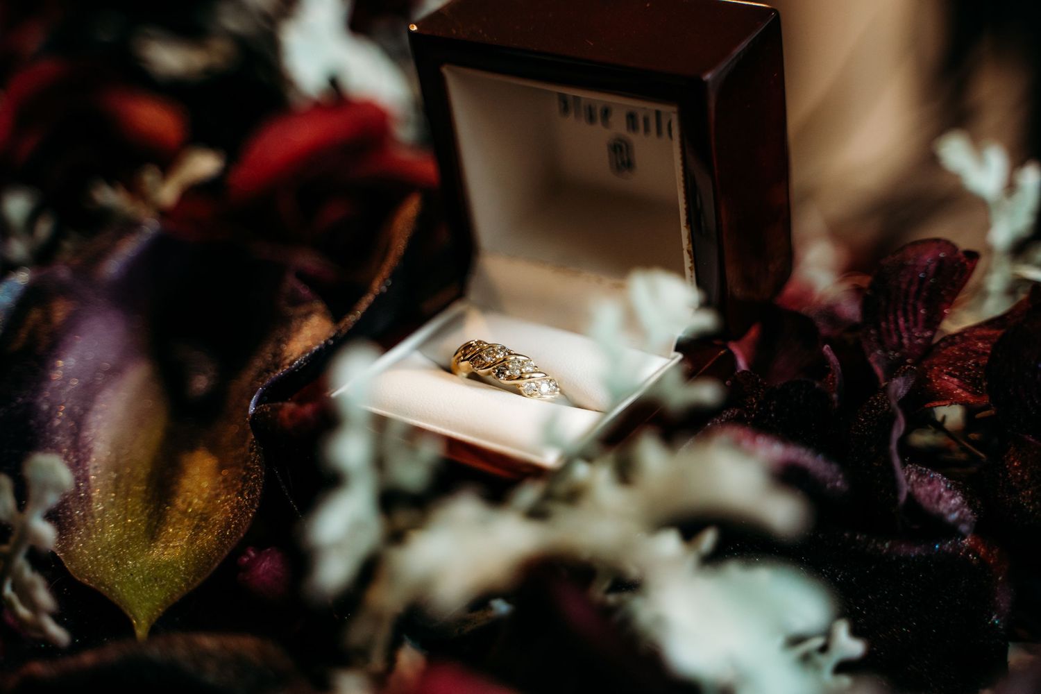 Close up of diamond engagement ring in jewelry box surrounded by dark purple flower petals.
