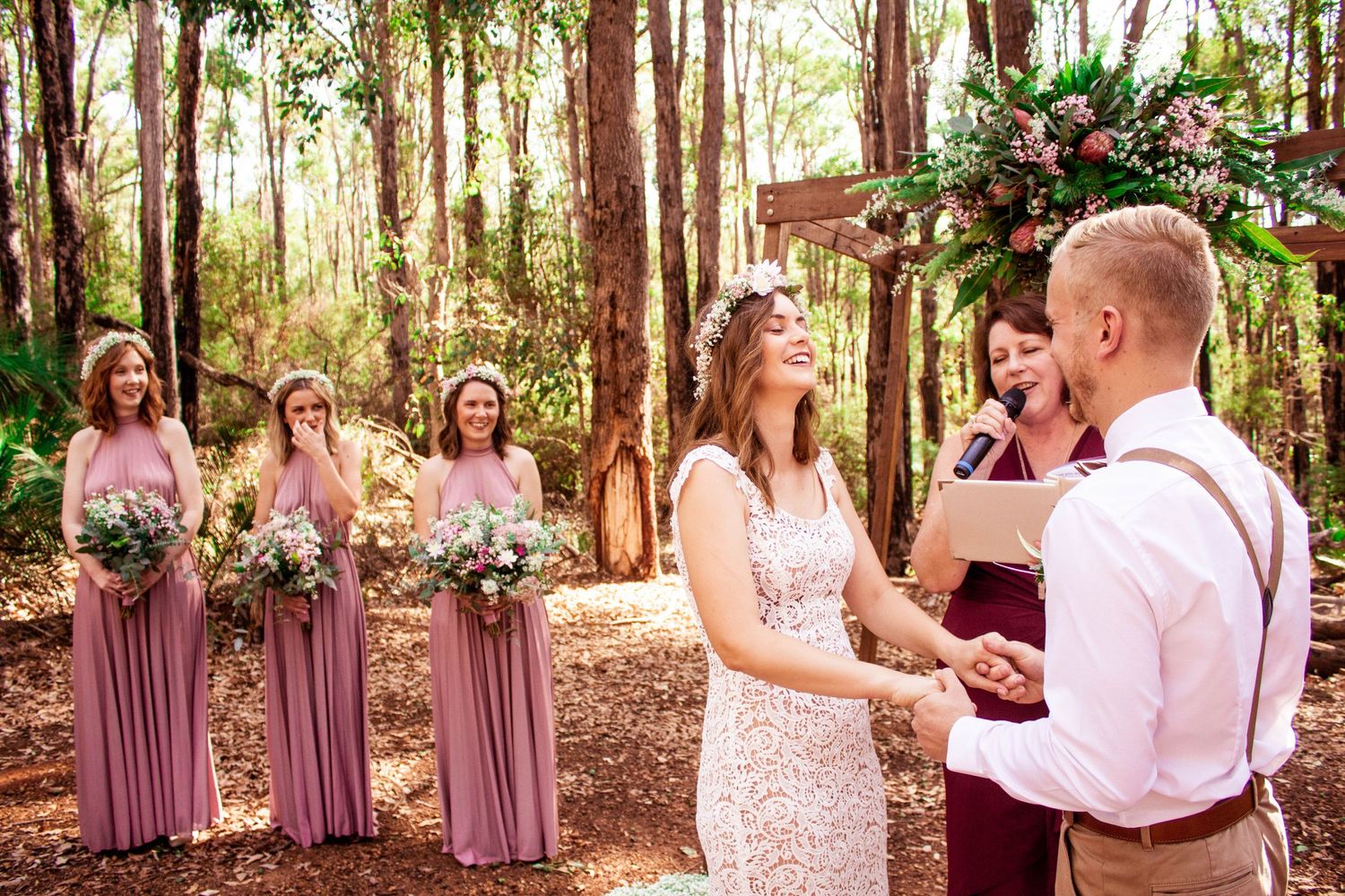 A woodland wedding ceremony with bridesmaids in mauve dresses watching the exchange of vows under tall trees.