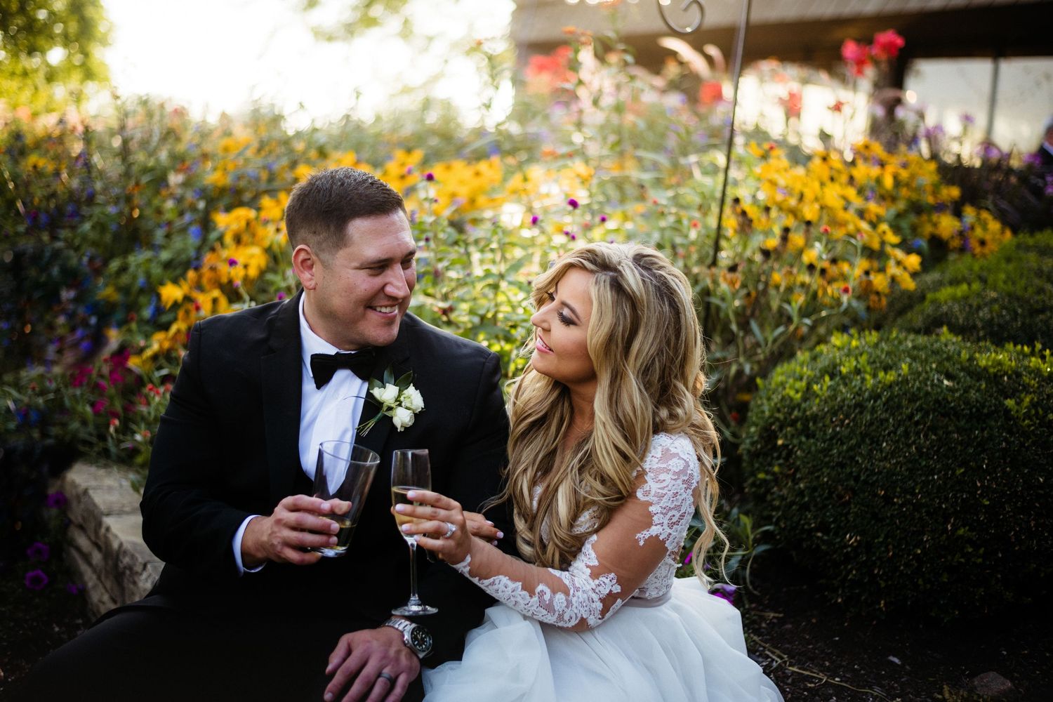 A couple toasts with wine glasses in a beautiful garden setting filled with vibrant yellow flowers at sunset.